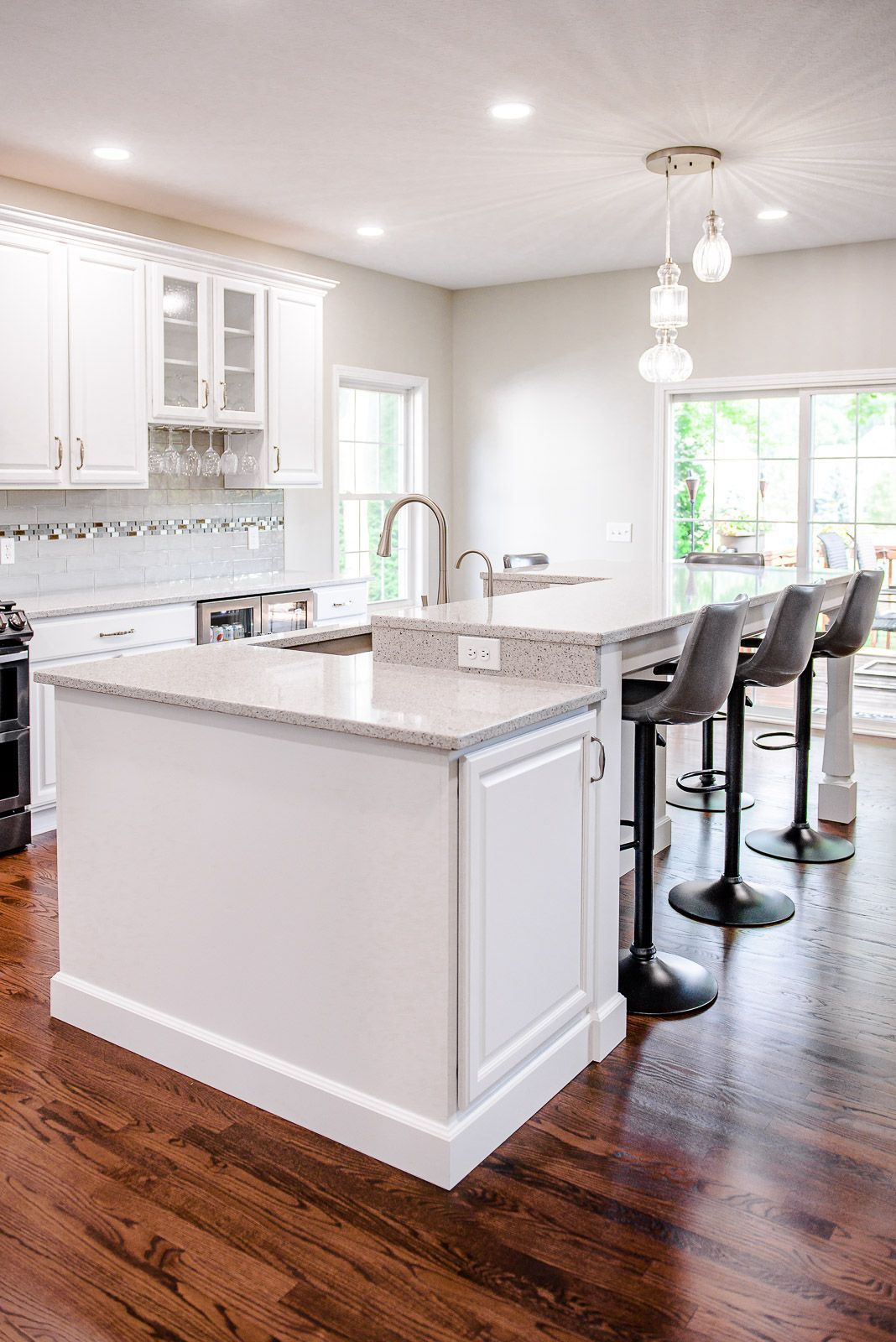 White kitchen with island, quartz countertops, wooden floor. Bar stools at the island, and a sink by the window.