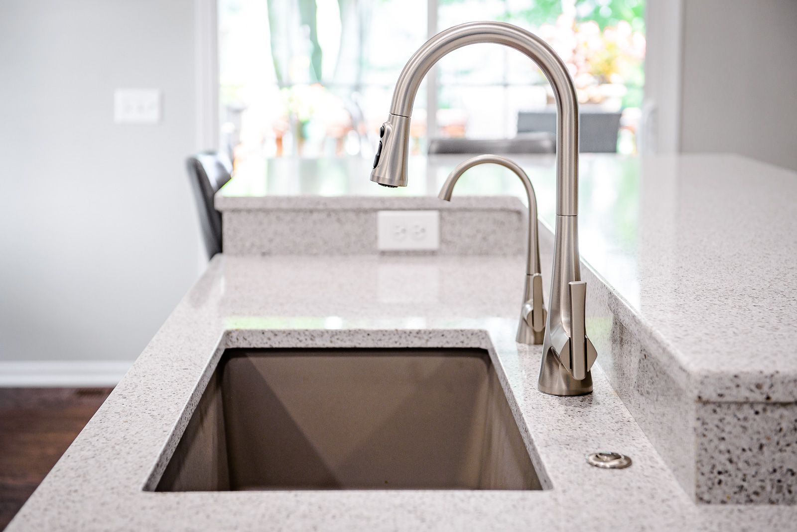 Stainless steel kitchen faucet over a square sink in a light speckled countertop.