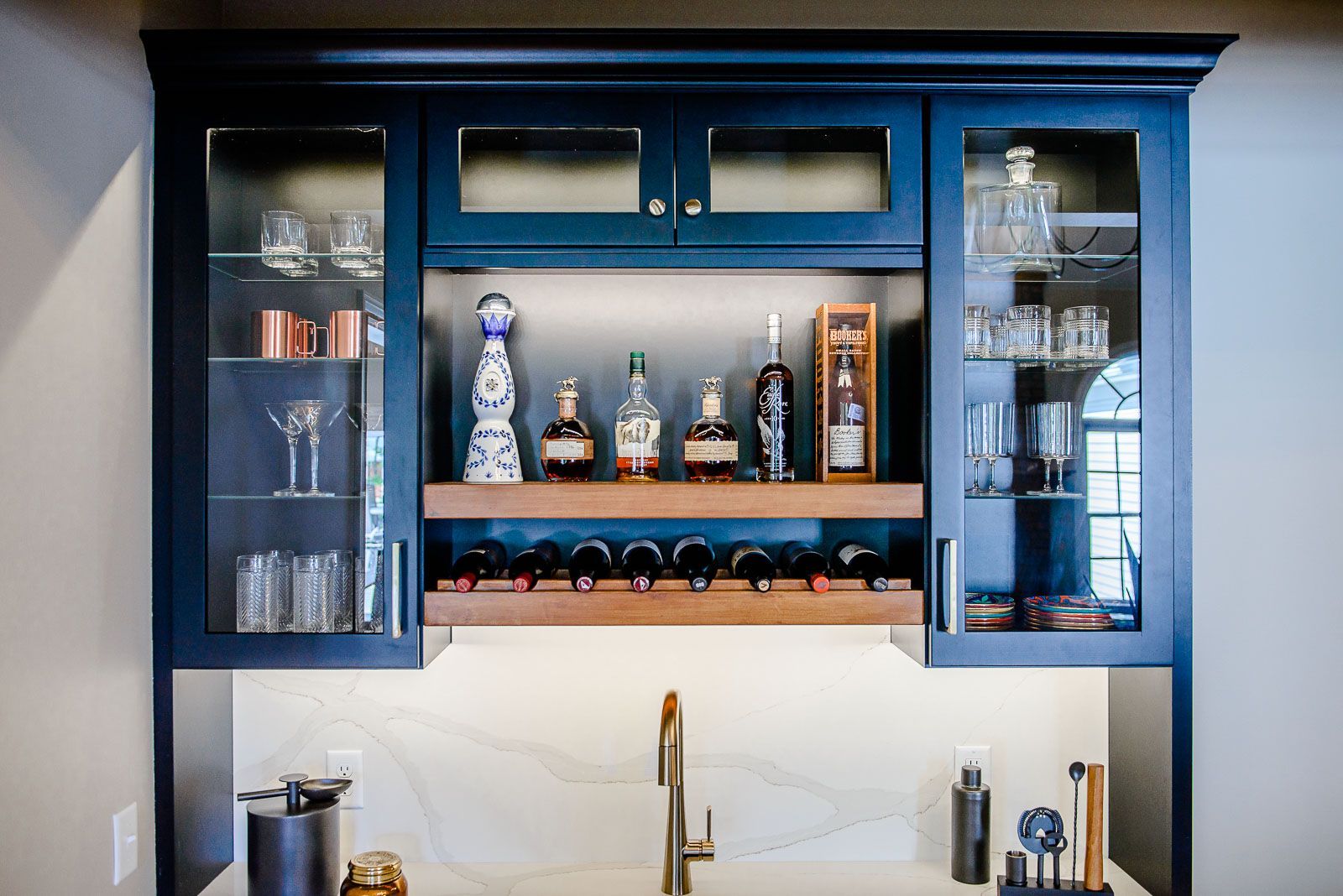 A kitchen with a sink , bottles , glasses and a wine rack.