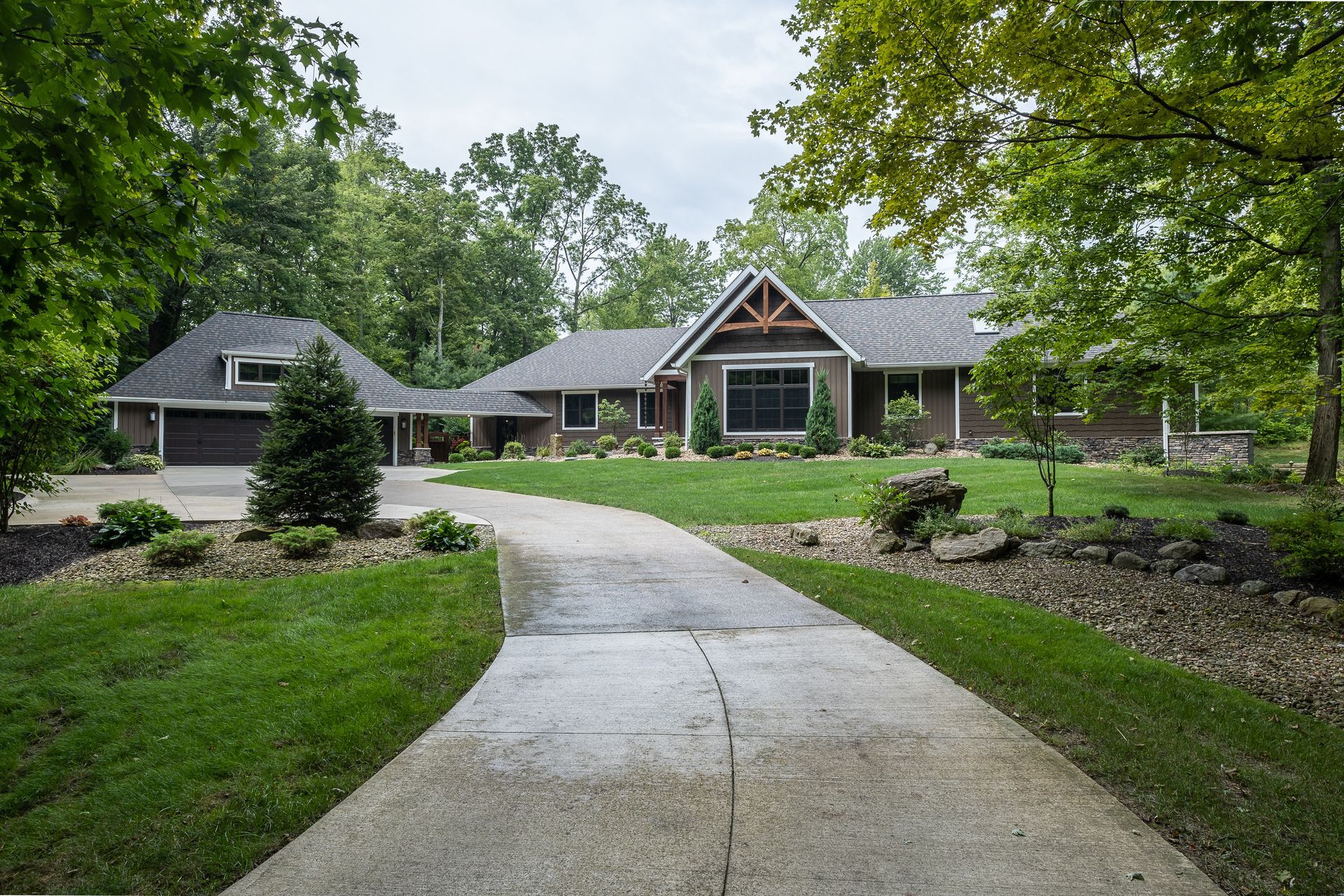 House with a long driveway surrounded by lush green lawn and trees. Brown siding and a two-car garage.