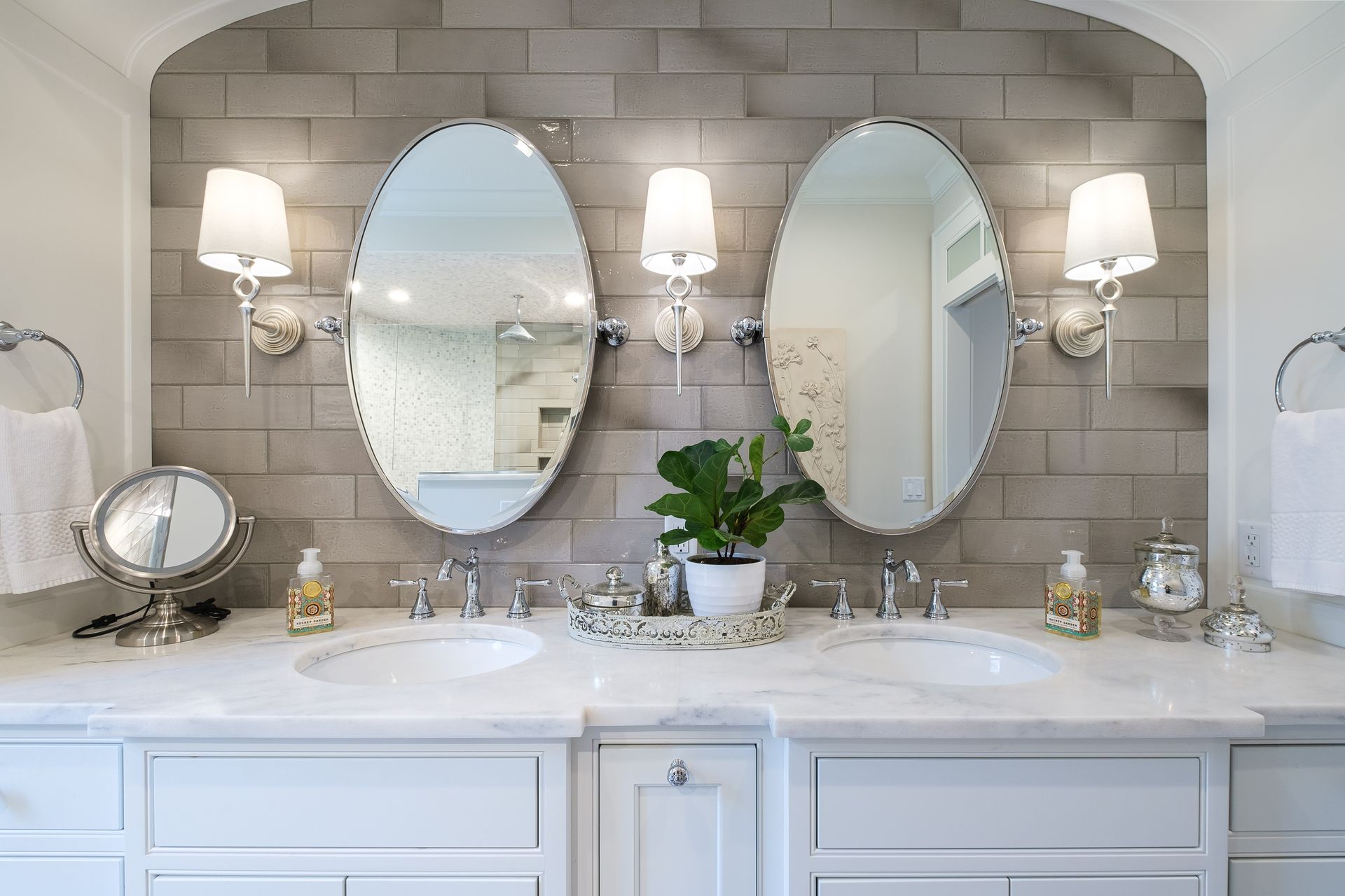 Bathroom with two oval mirrors, gray brick backsplash, white sinks, and bright sconces.