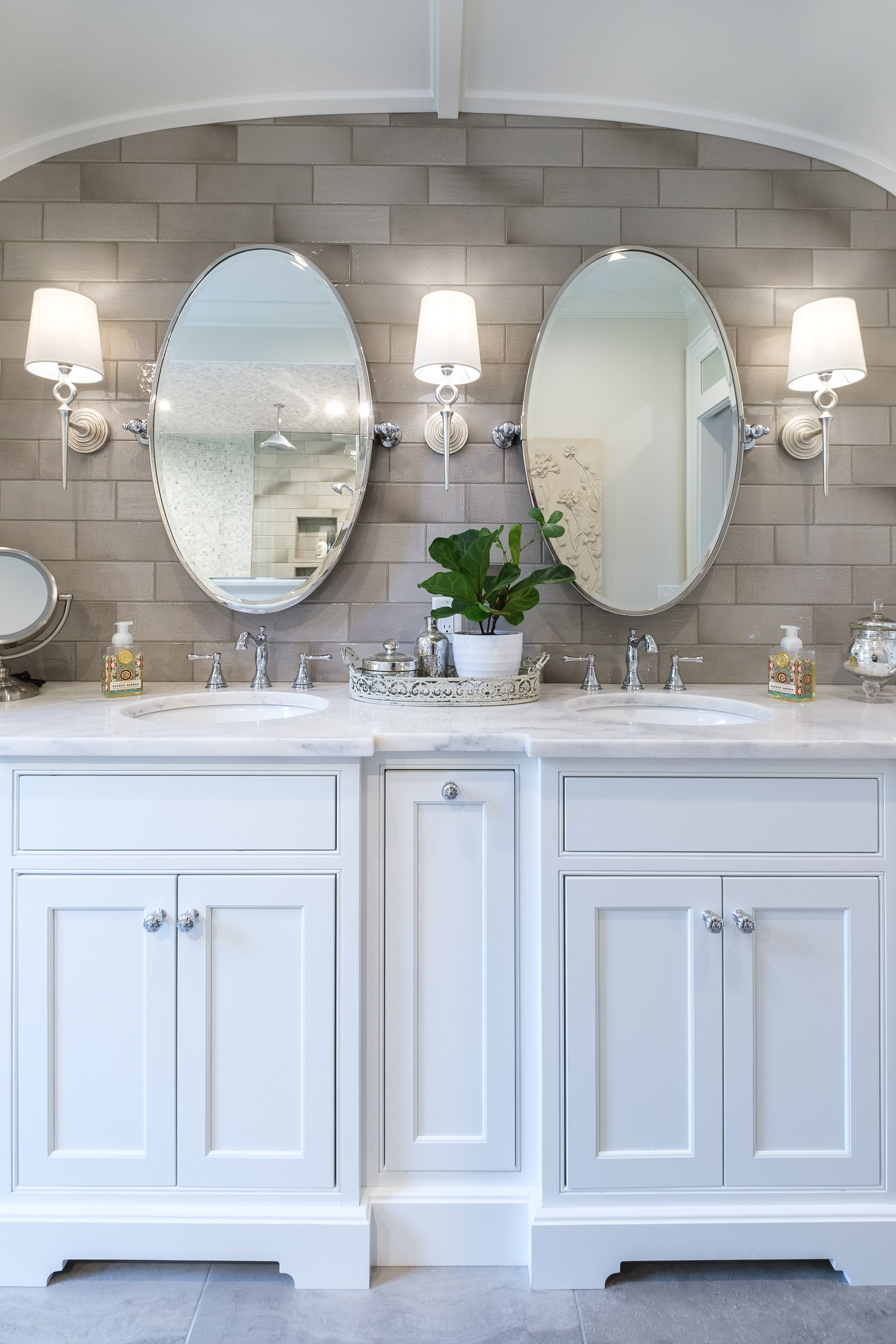 White bathroom vanity with two oval mirrors, sconces, and a plant against a gray tiled wall.