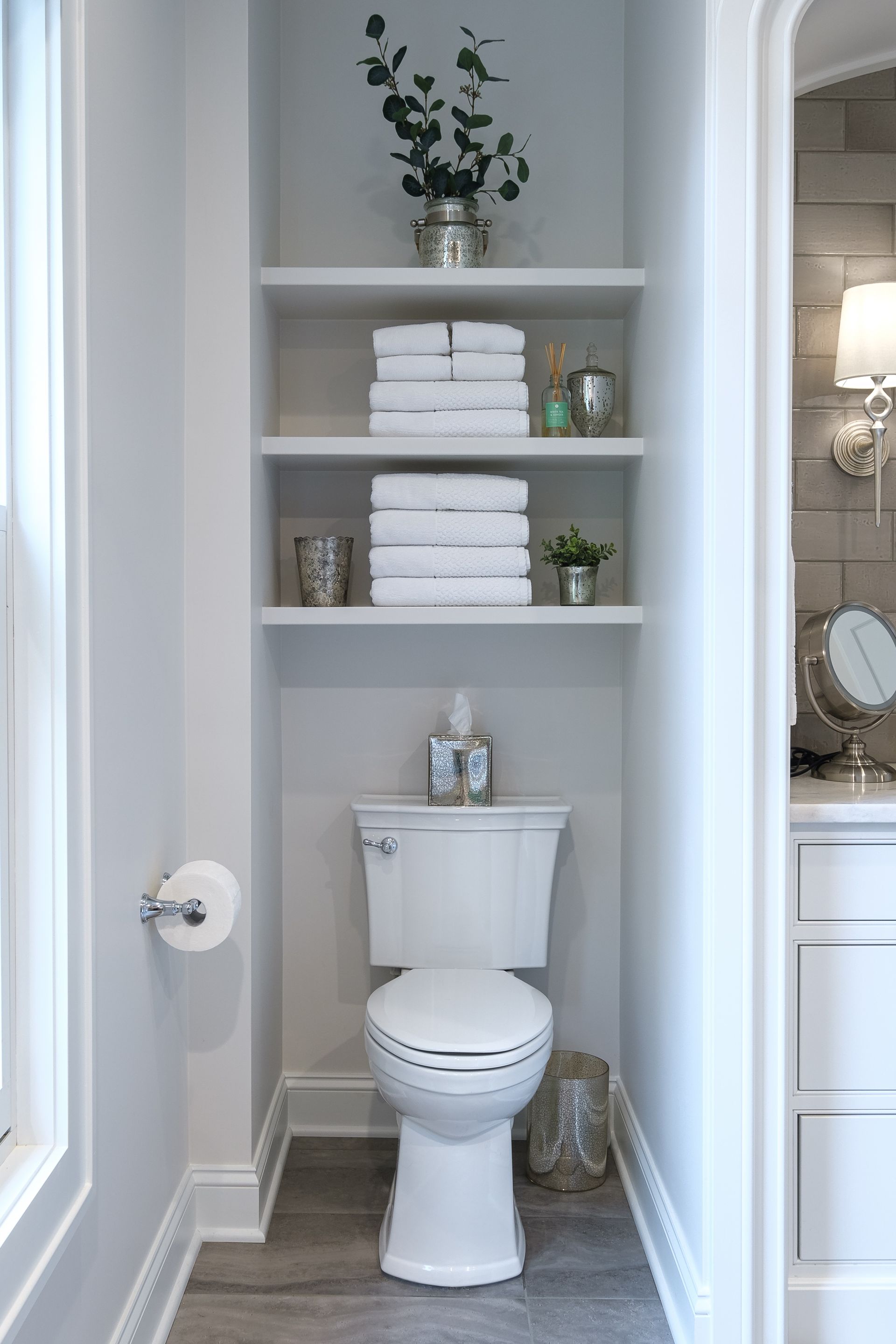 Narrow bathroom with toilet beneath built-in shelves holding towels, plants, and decor.