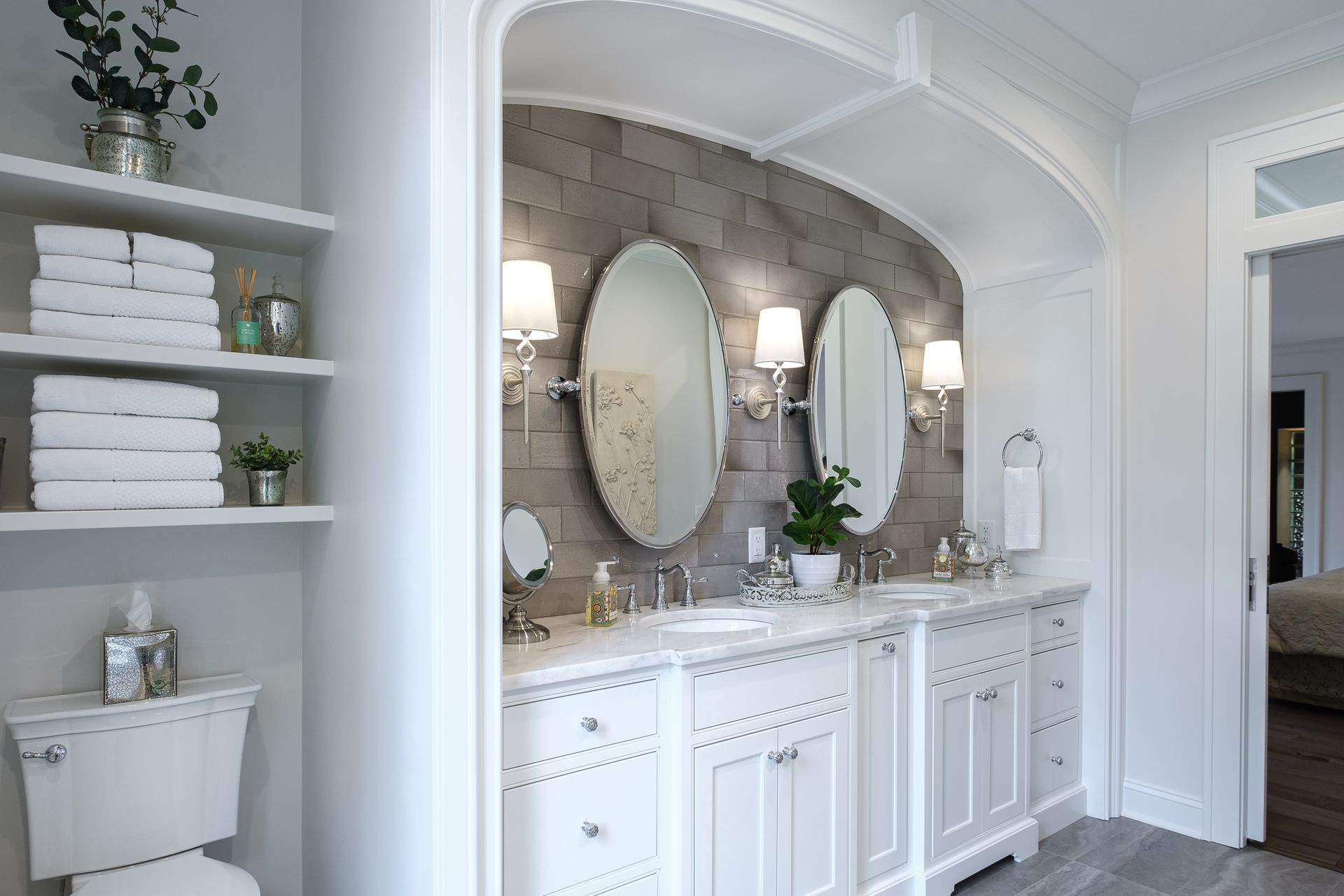 Elegant white bathroom with double vanity, oval mirrors, and gray brick wall accent.