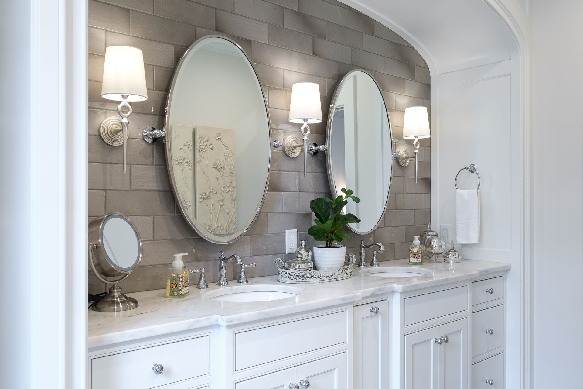 Bathroom with white vanity, oval mirrors, gray brick wall, sconces, and a plant.