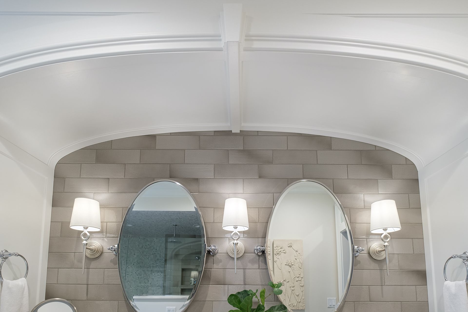 Bathroom vanity with oval mirrors, sconces, and gray tiled wall, under a white arched ceiling.