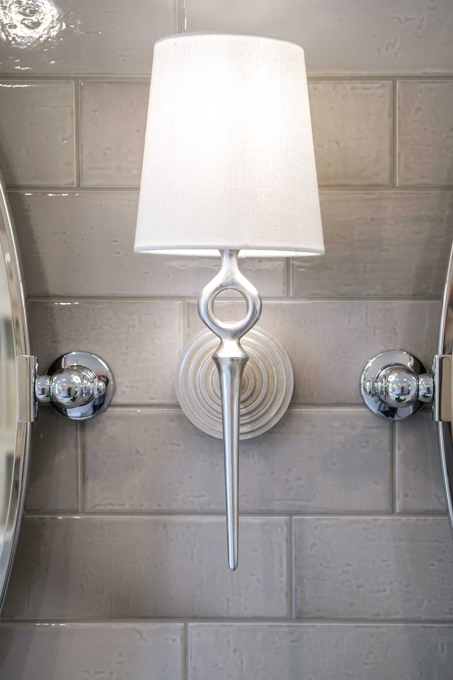 Silver sconce lamp with white shade, between two chrome-rimmed mirrors on gray tile wall.