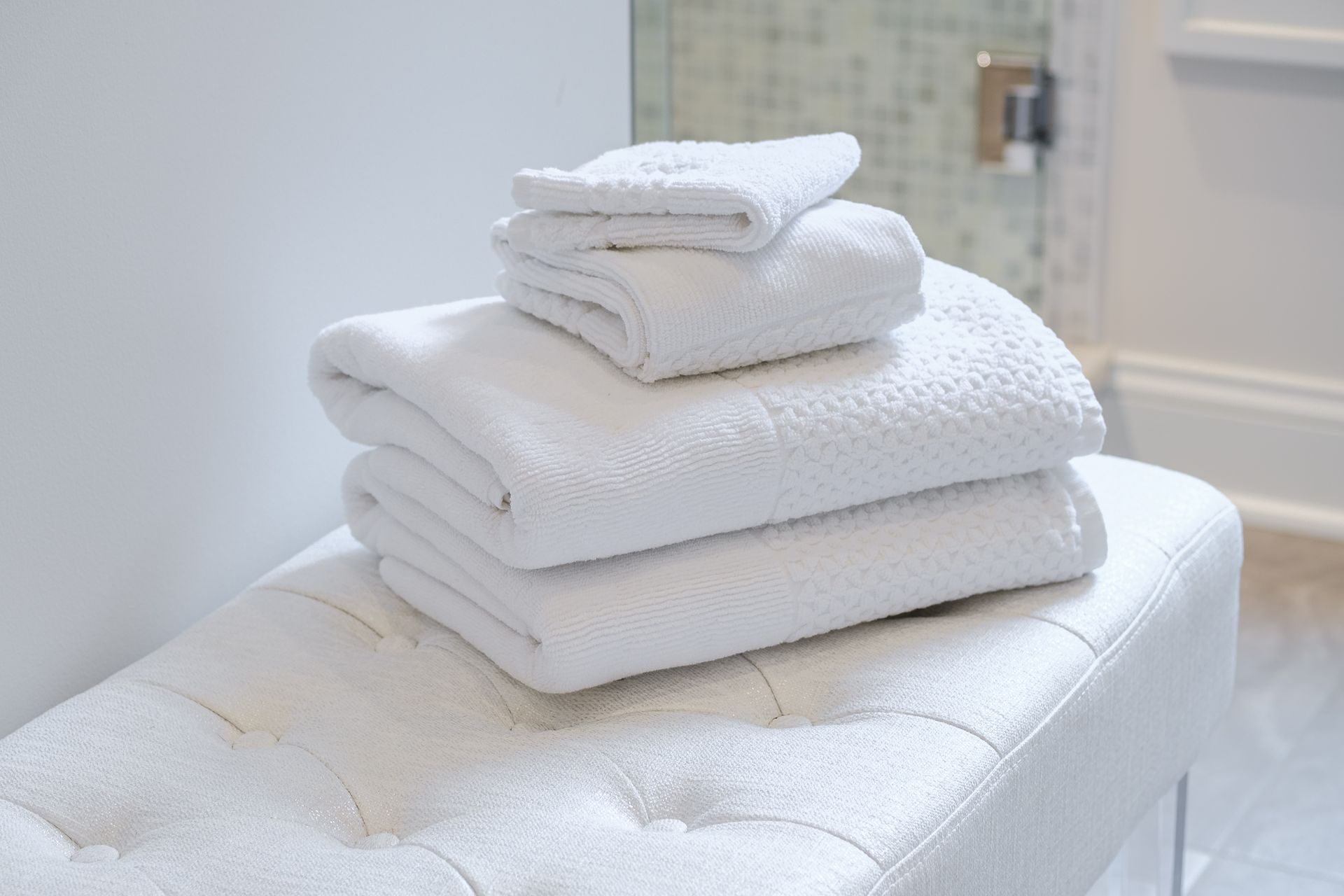 White towels stacked on a tufted bench in a bright bathroom.