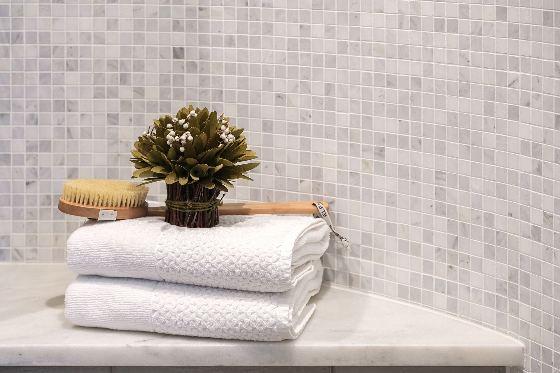 White towels, brush, and plant on marble shelf in front of white mosaic tile wall.