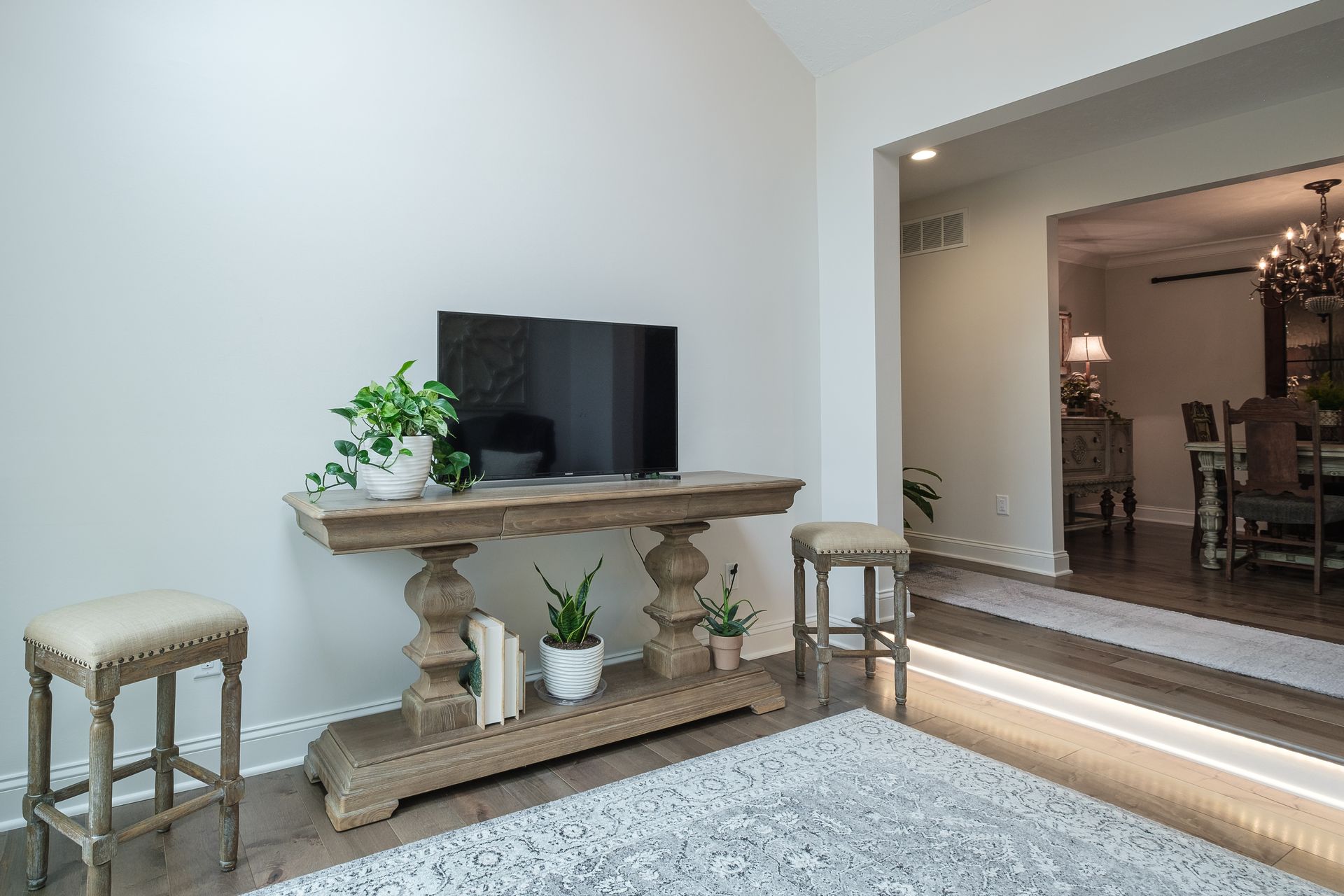 A living room with a TV on a wooden console, plants, stools, and a rug. Open doorway to a dining room.