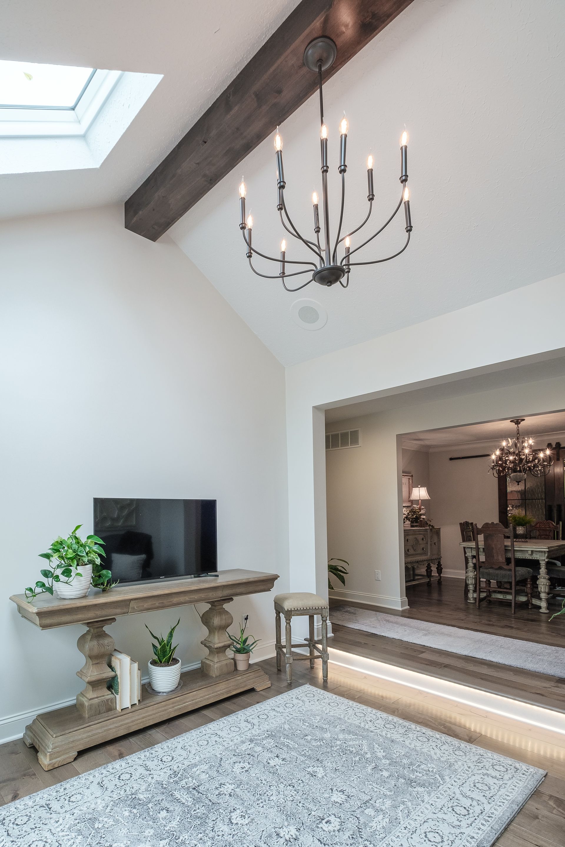 Living room: white walls, wood beam, chandelier, TV on console table, rug, view into dining room.