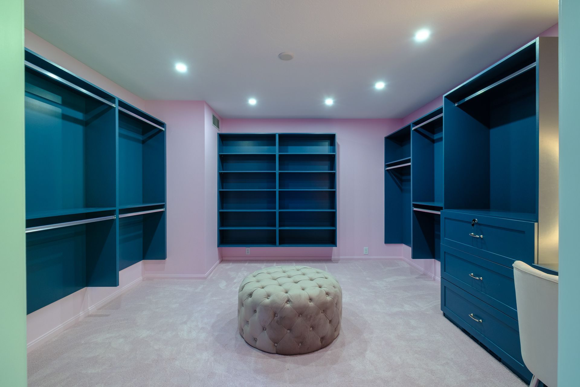 Pink and blue walk-in closet with shelves and drawers; a tufted ottoman sits in the center.