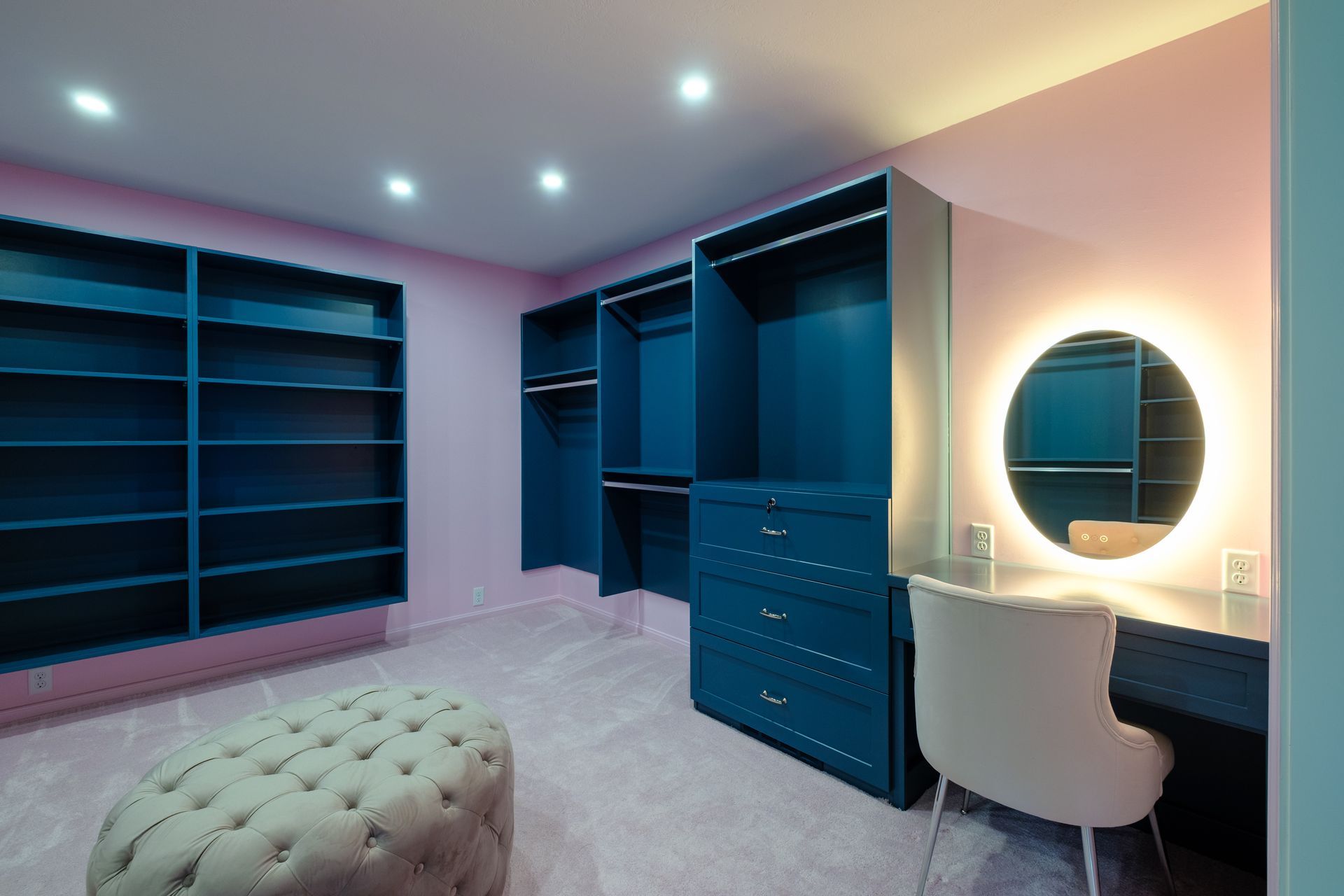 A walk-in closet with blue cabinetry, pink walls, a vanity with a circular mirror, and an ottoman.