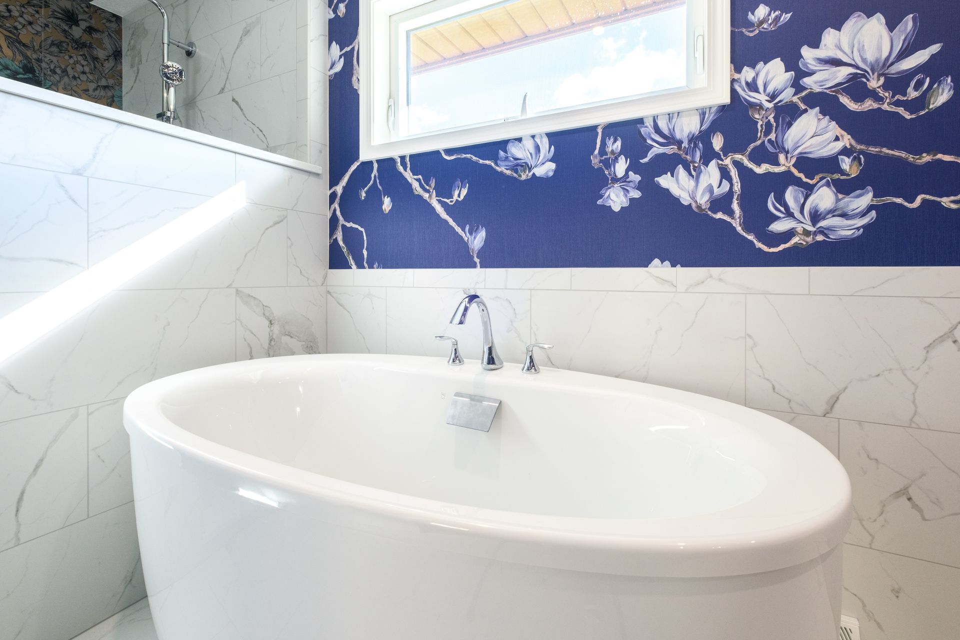 Oval bathtub in a bathroom with blue floral wallpaper, marble tile, and a window.