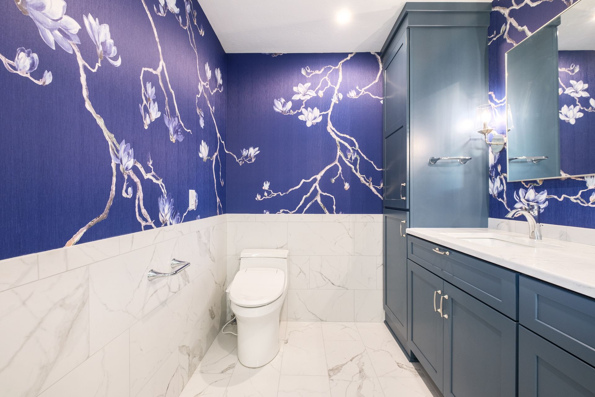 Blue floral wallpaper bathroom with white marble tile, blue cabinets, and white toilet.