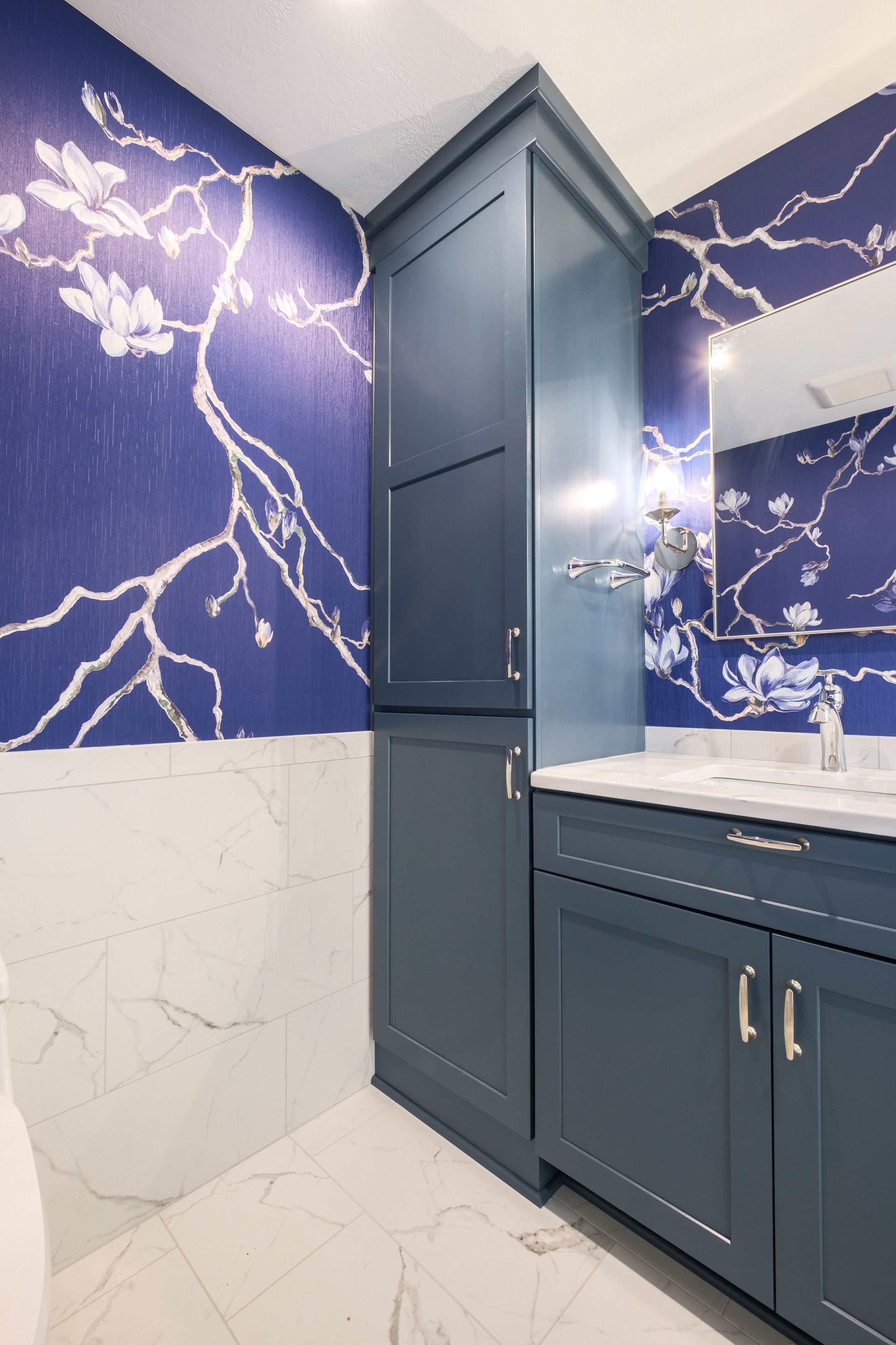 Blue and white floral wallpaper, blue cabinets, marble countertop in a bathroom.