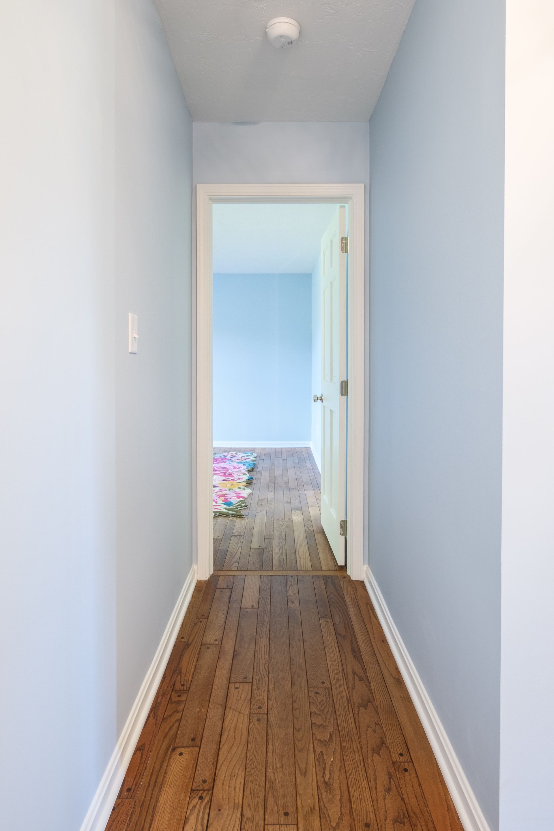Narrow hallway with light blue walls, wooden floor, and a door at the end leading to a blue room.
