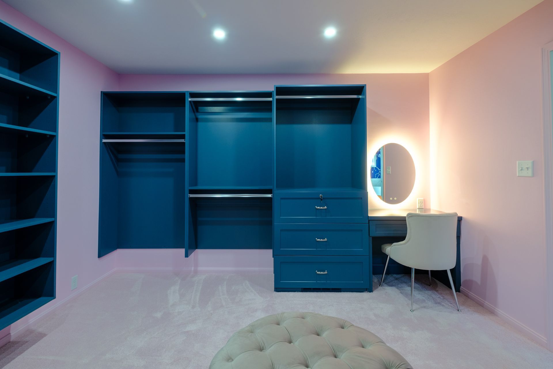 Walk-in closet with blue cabinetry, a vanity, and pink walls. A neutral ottoman sits on the carpet floor.