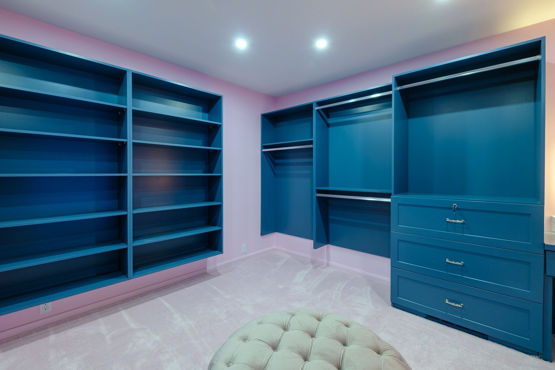 Blue closet shelving against pink walls and carpet; a tufted ottoman in the foreground.