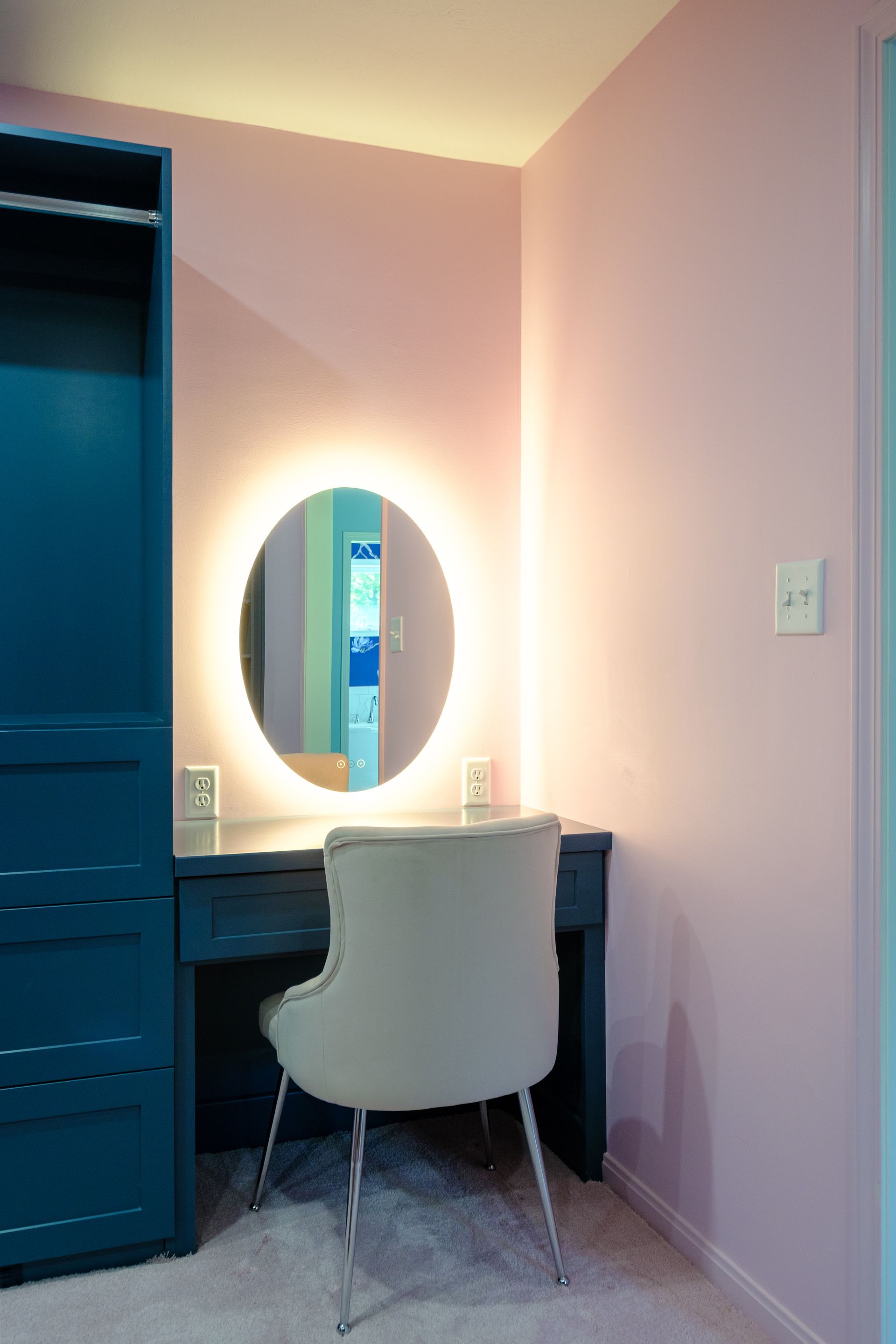Vanity area with oval lighted mirror, teal vanity and closet, pink walls, and grey chair.