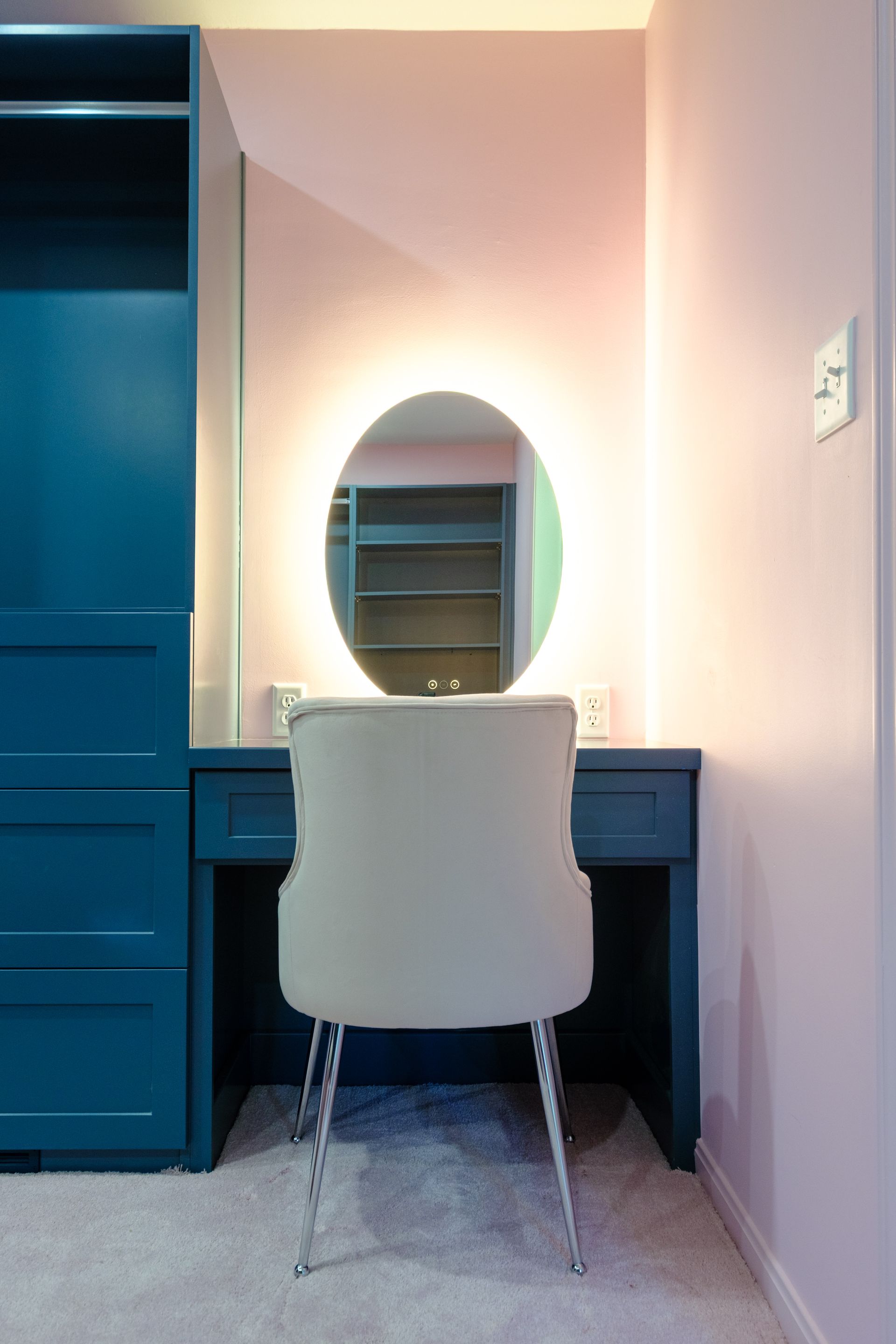 Vanity with oval, backlit mirror, gray chair, teal cabinet, and pink wall.