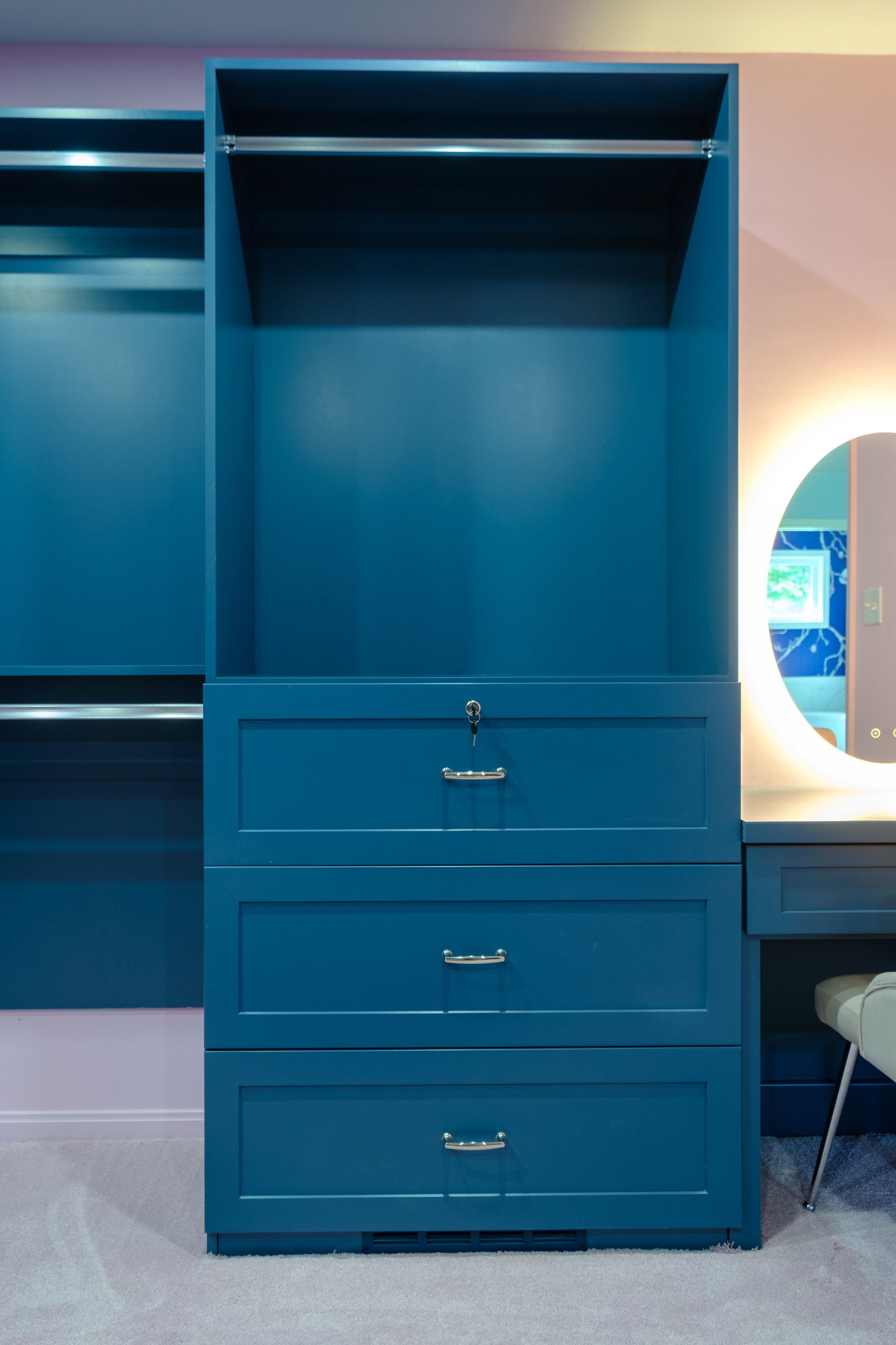 Blue wardrobe with drawers and hanging rod, next to a vanity mirror in a bedroom.