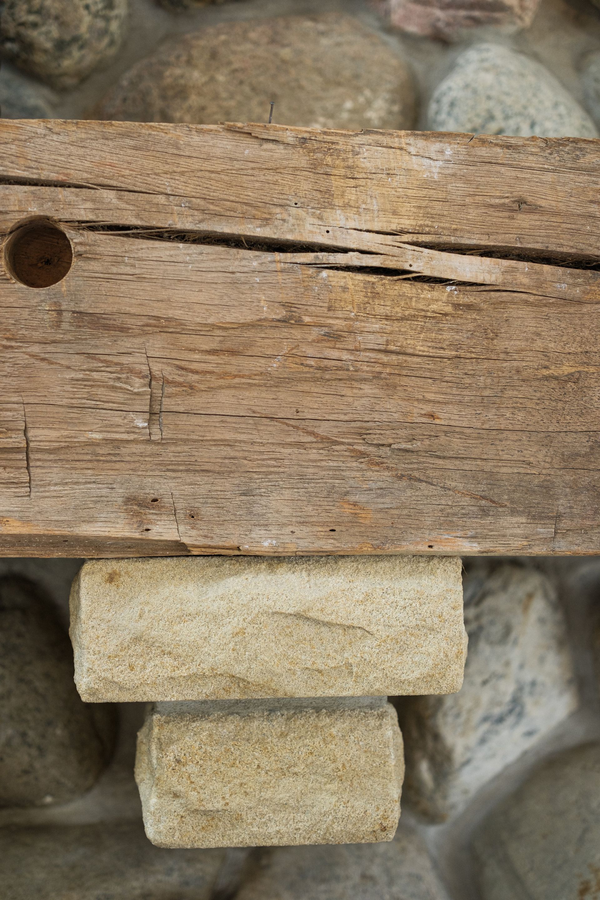 Wooden beam resting on two light-colored stone blocks, set against a background of river stones.