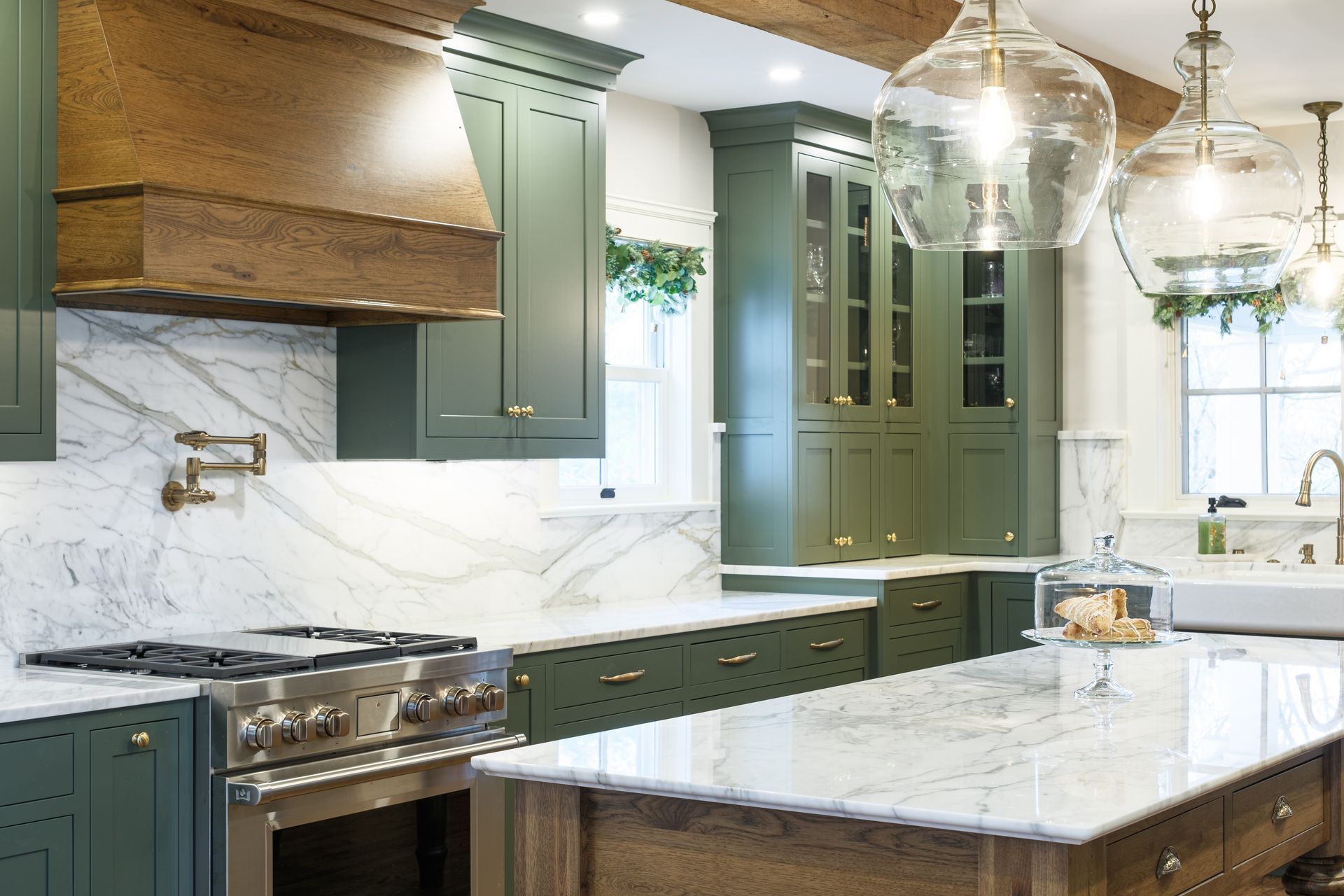 Green kitchen with marble countertops and a stainless steel range, lit by pendant lights.