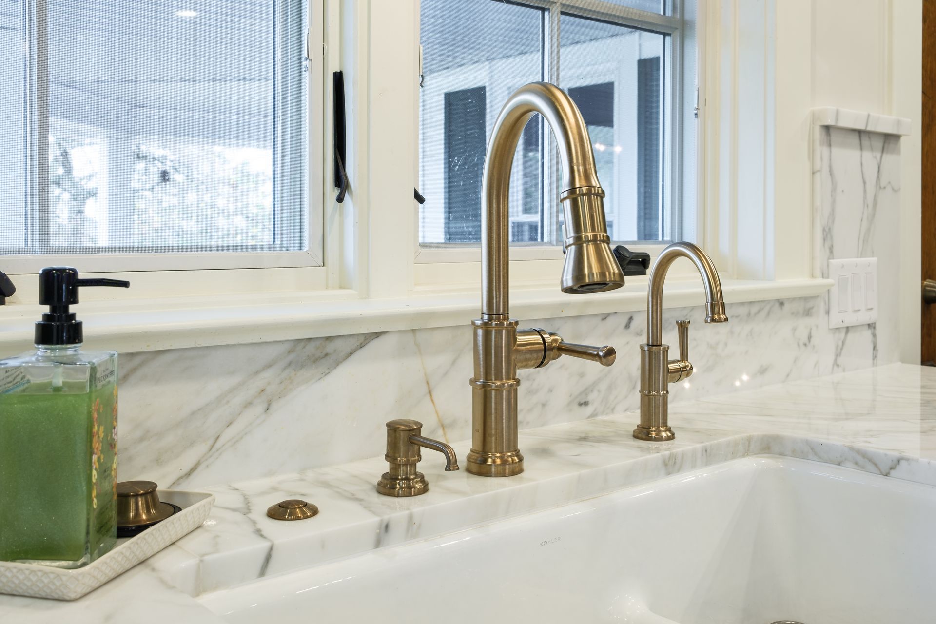Golden kitchen faucet and soap dispenser on a marble countertop with a window in the background.
