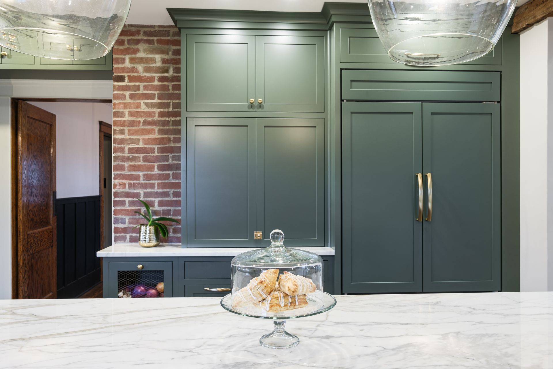 Kitchen with sage green cabinets, brick wall, and marble countertop. Glass cloche with pastries.