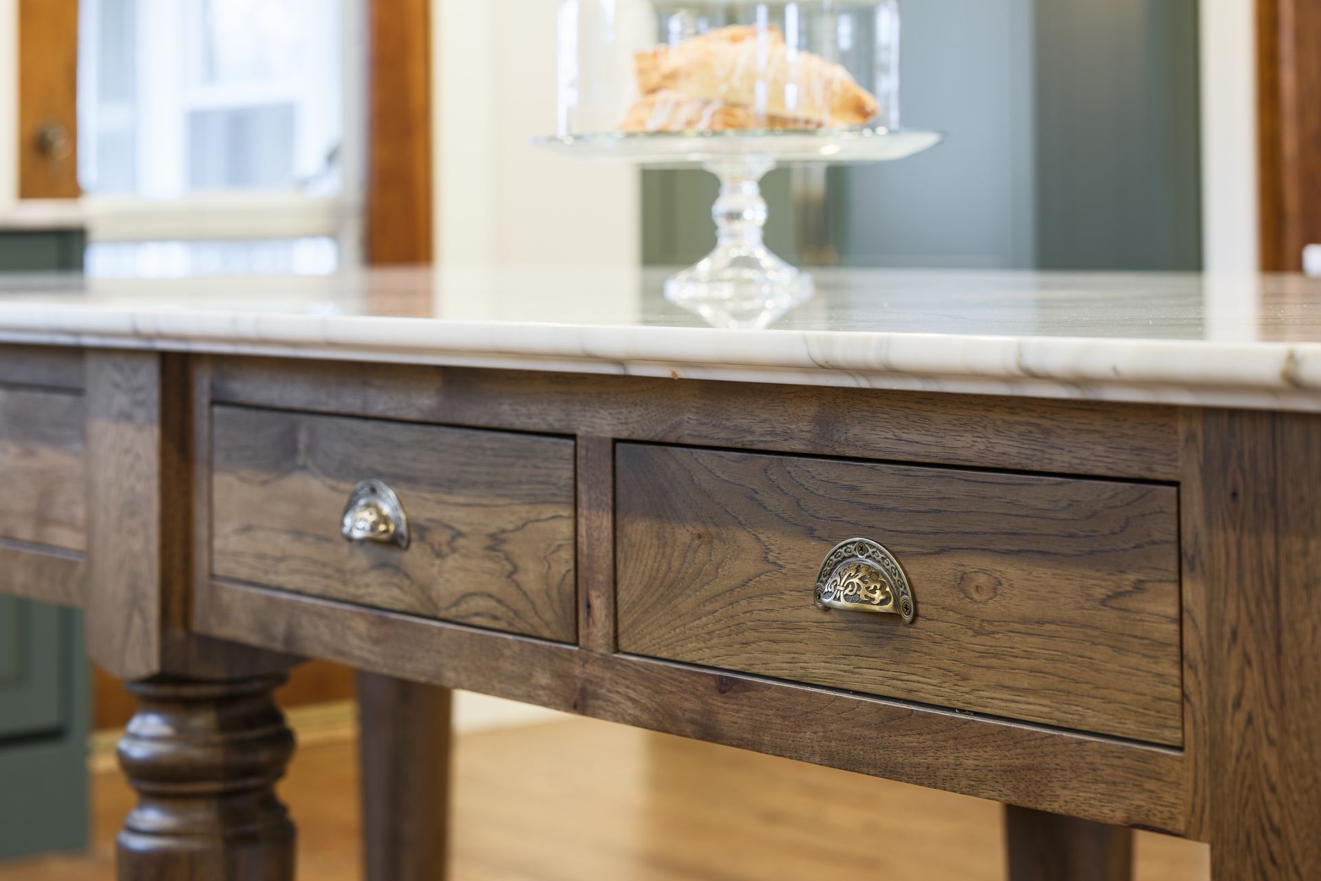 Wooden kitchen island with marble countertop, two drawers with brass handles, and glass cake dome.