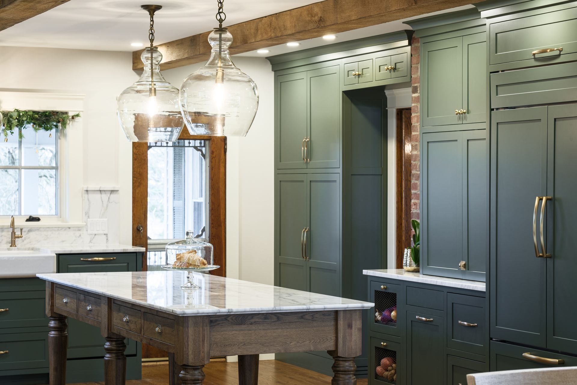 Green kitchen with island, marble countertop, glass pendant lights, and wooden door.