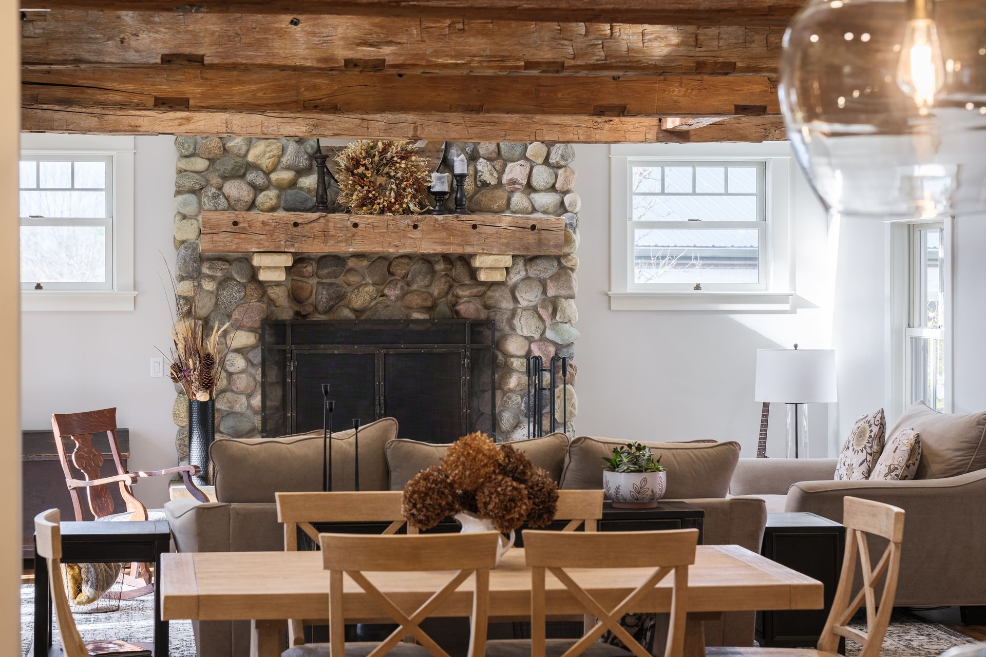 Rustic living room with stone fireplace, wooden beams, dining table, and two seating areas.