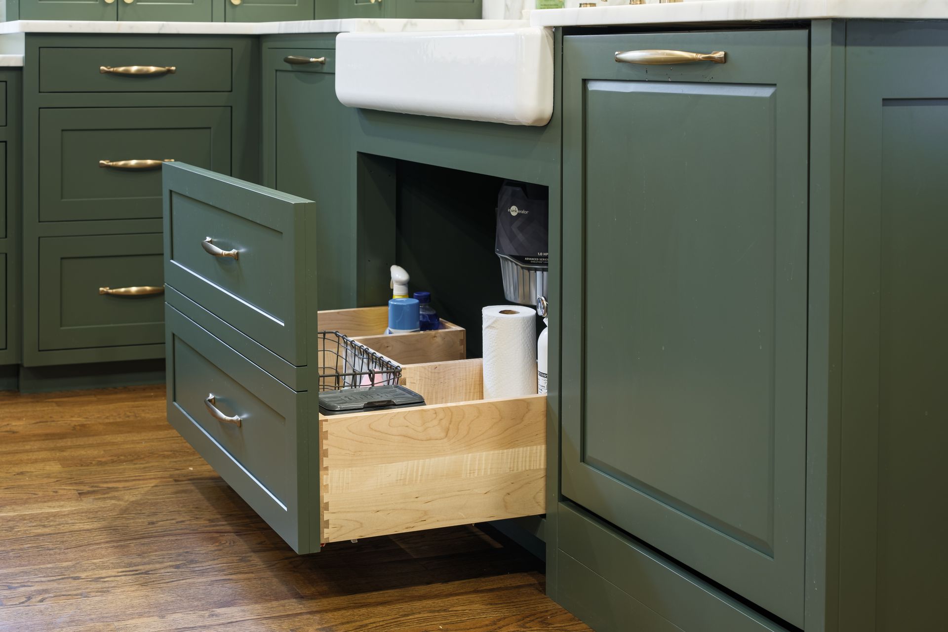 Green kitchen cabinets with a drawer open, revealing cleaning supplies, under a white farmhouse sink.