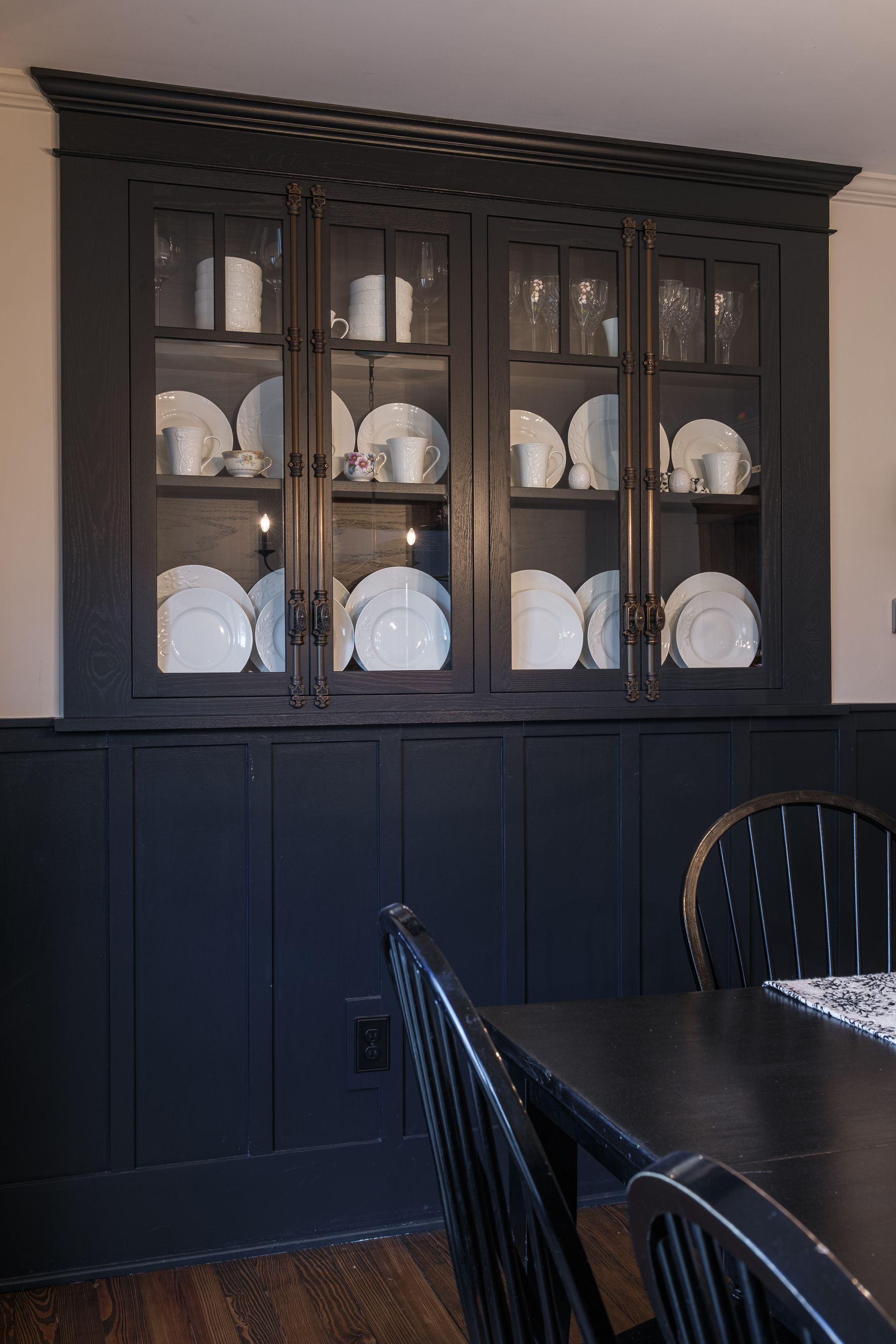 Black hutch with glass doors displays white dishes; set against black wainscoting.