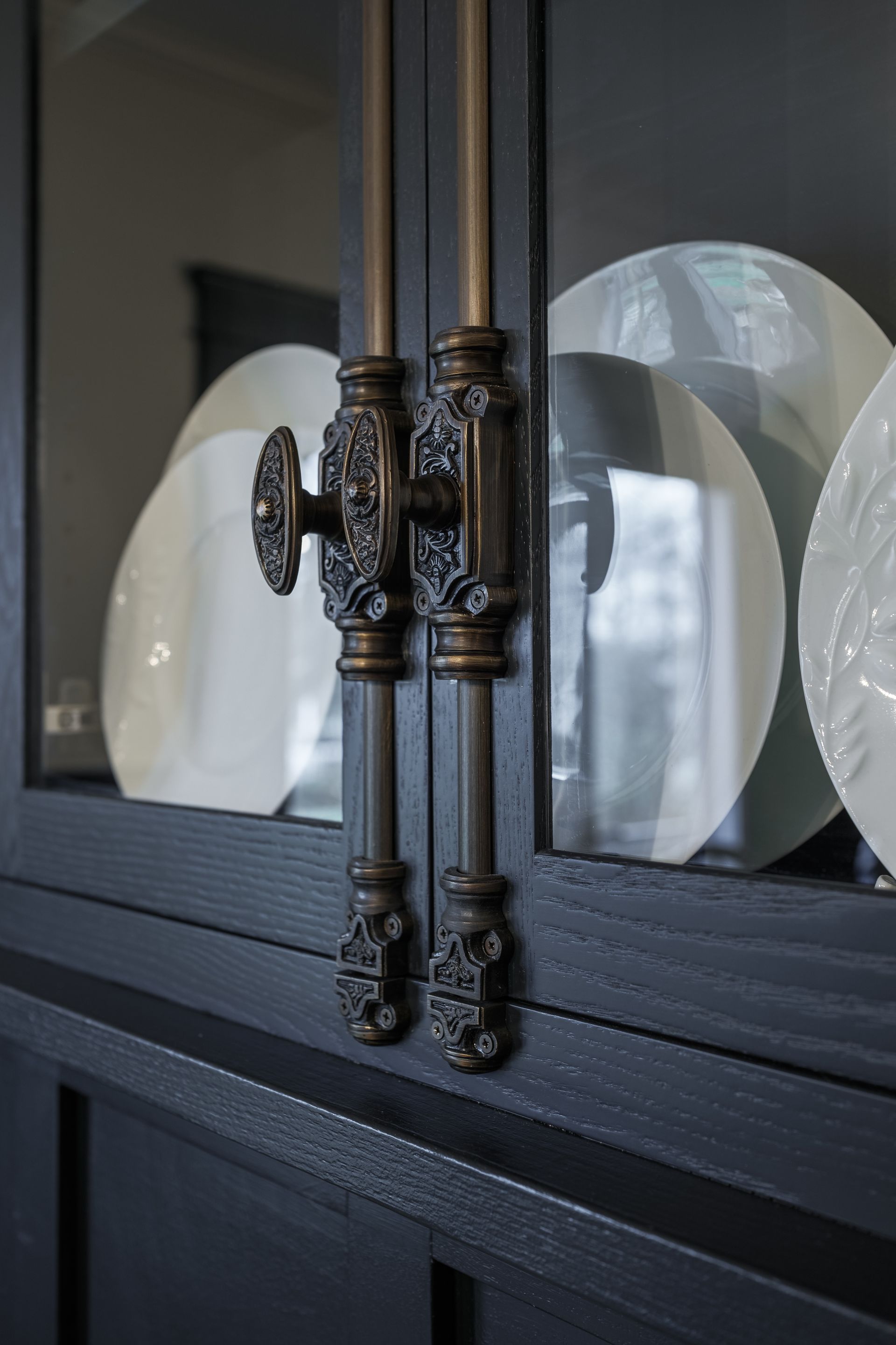 Close-up of a dark cabinet with ornate bronze handles and a decorative knob, displaying white plates.