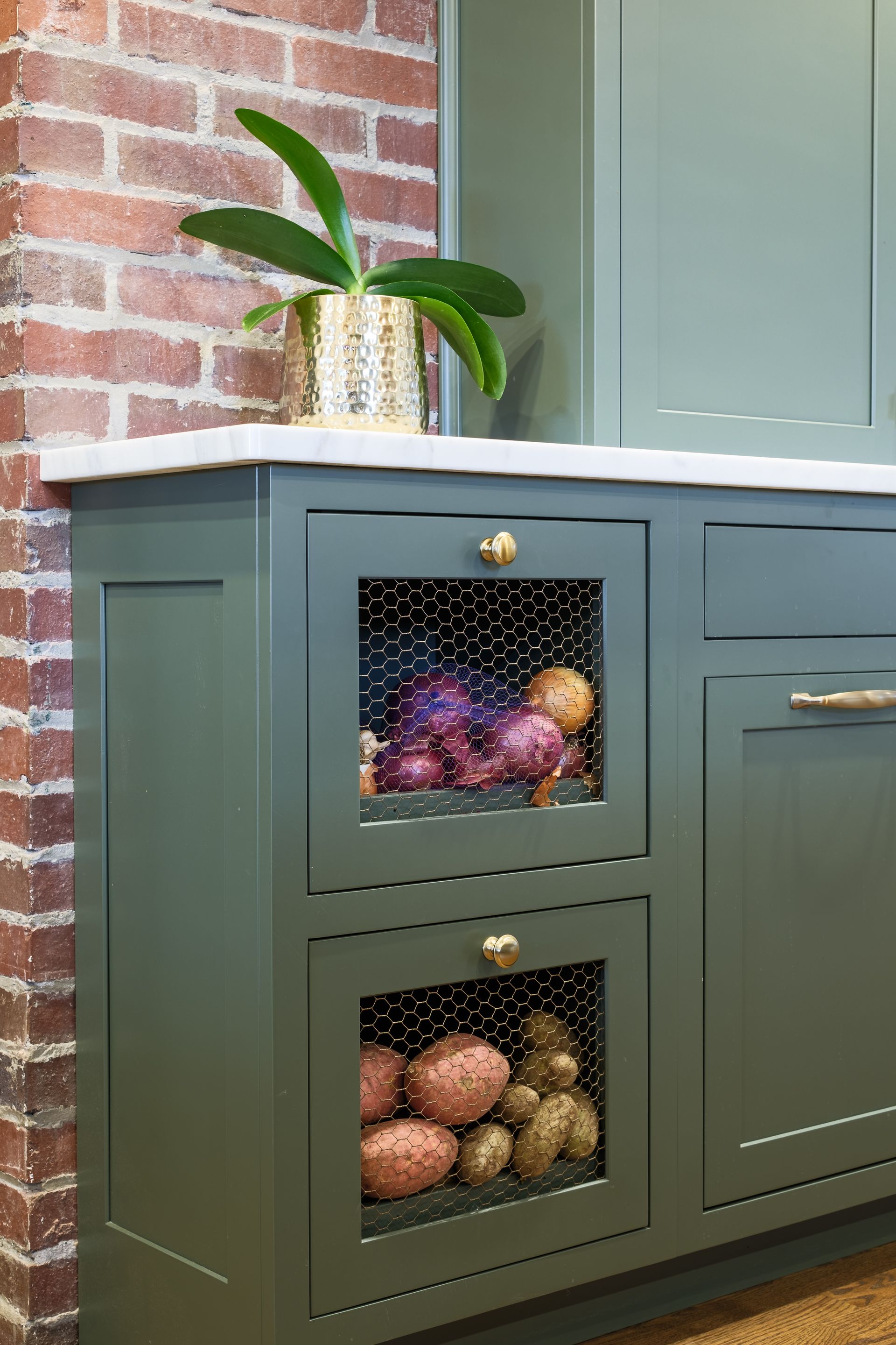 Green kitchen cabinet with wire mesh doors, holding potatoes and onions. Gold knobs and brick wall.