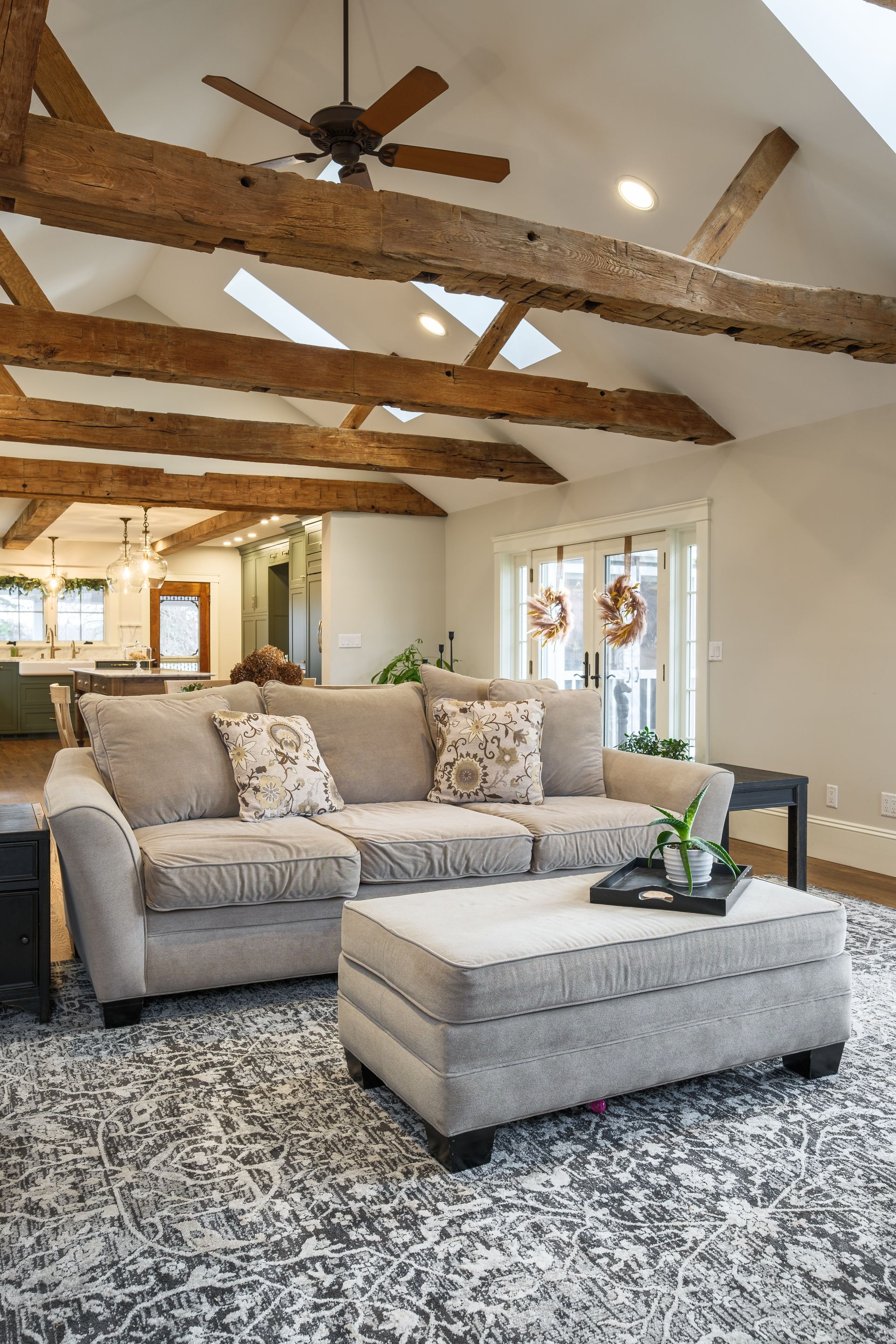 Living room with beige sofa, ottoman, patterned rug, wooden beams, and a view into the kitchen.
