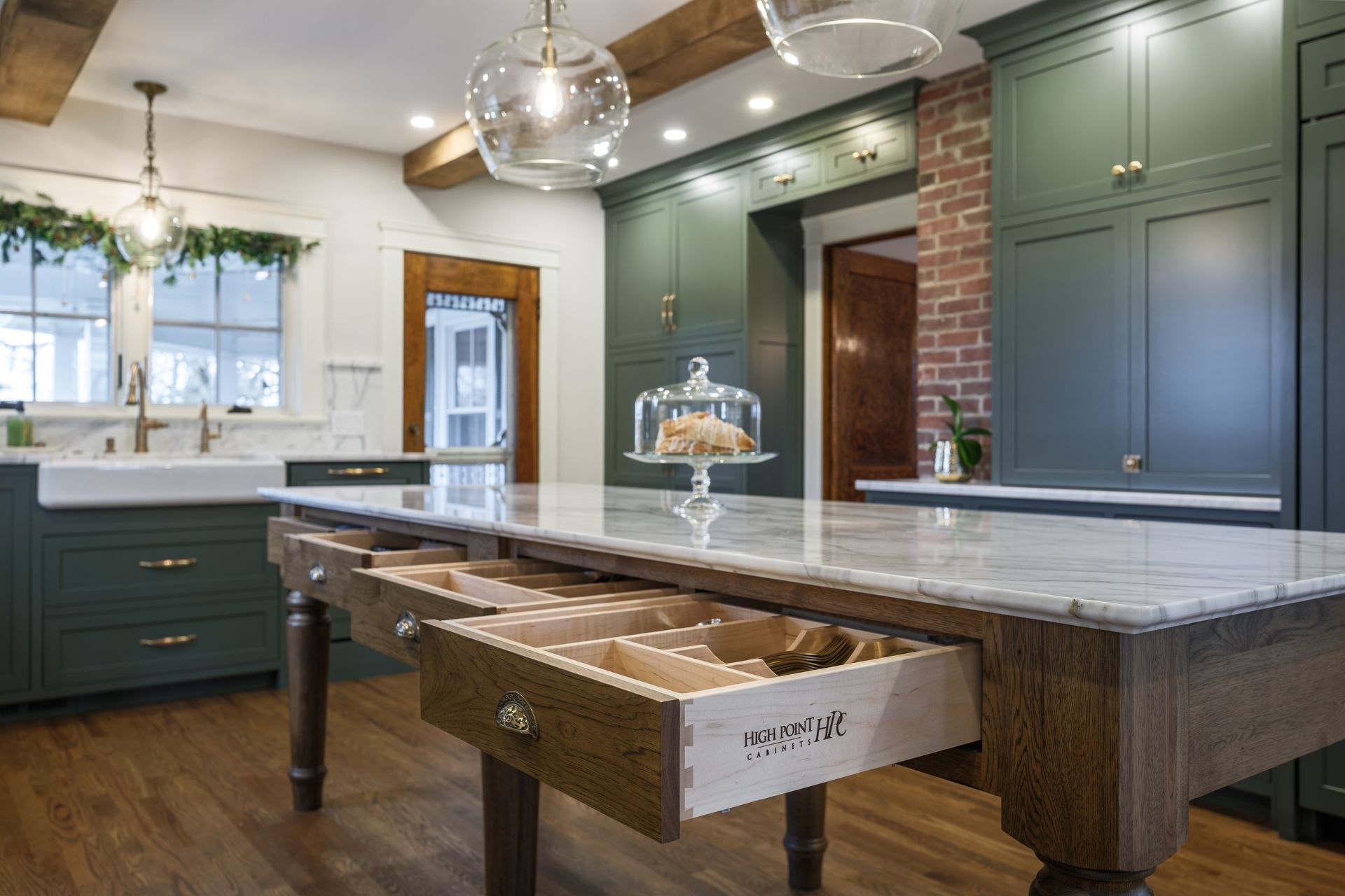 Green kitchen with a marble-topped island and open drawers, brick wall, and decorative glass lights.