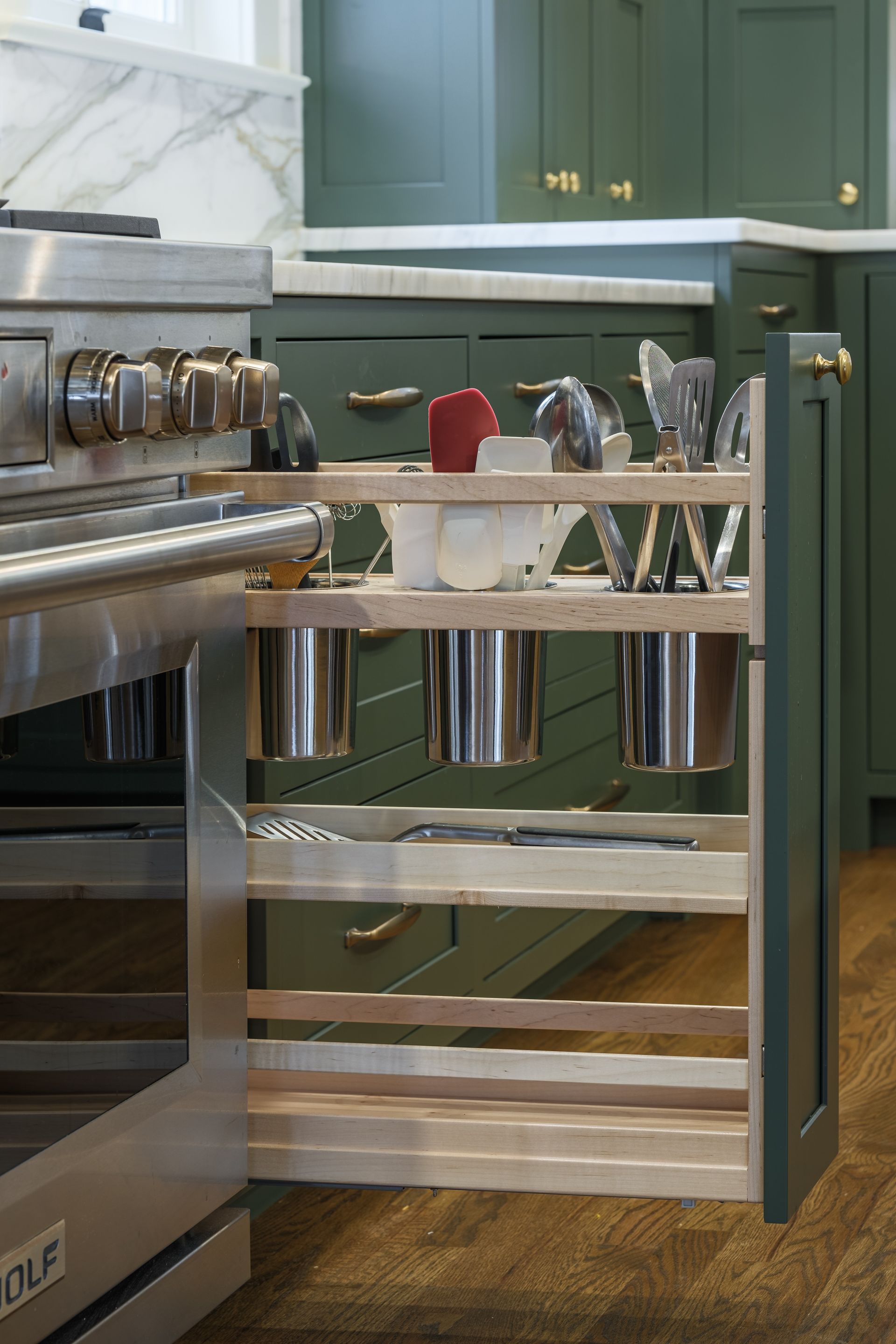 Open drawer beside a stove with utensils in metal containers. Green cabinets and wood floor.