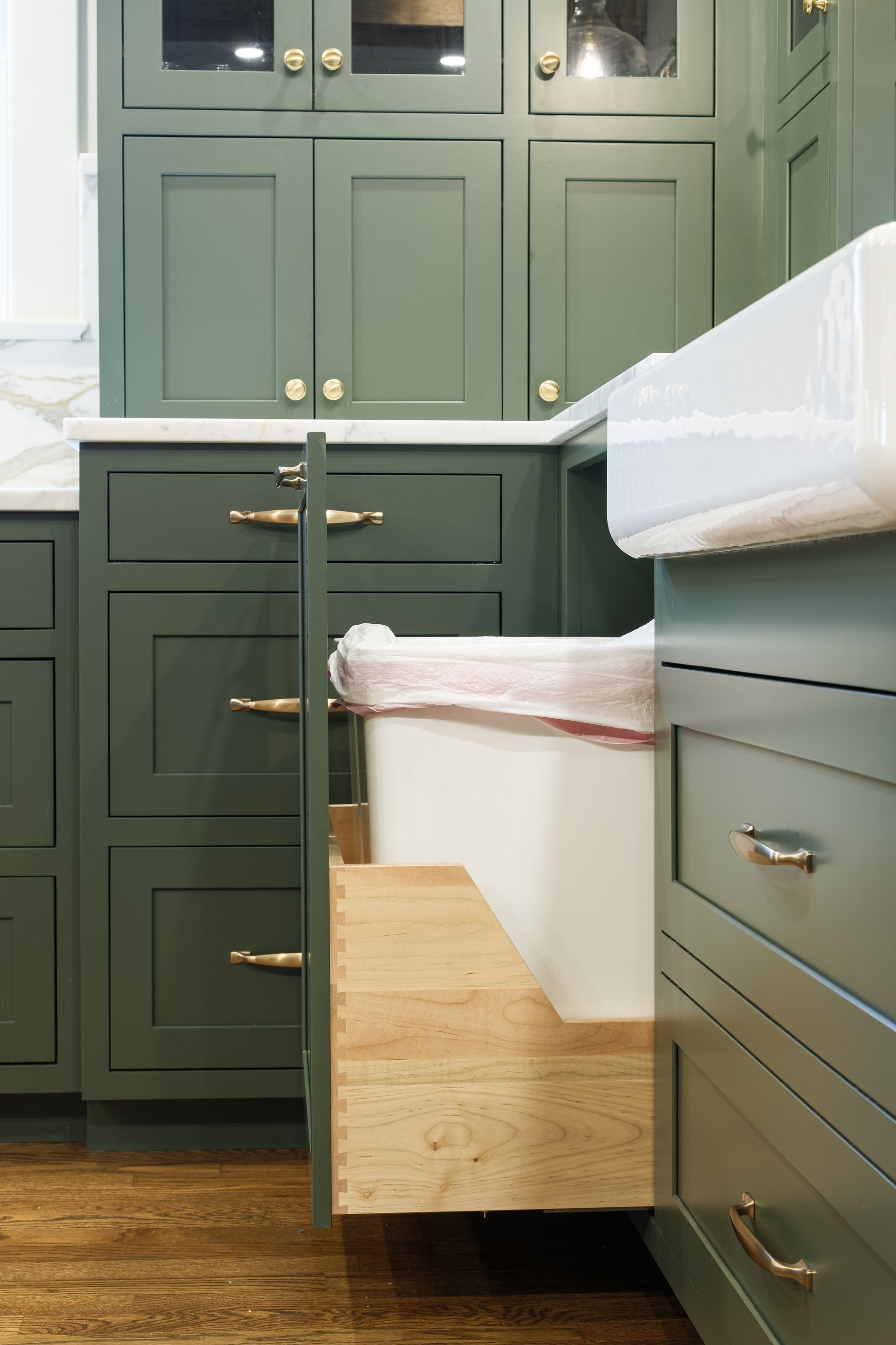 Green kitchen cabinets with a pull-out trash can. Gold hardware, wooden floors.