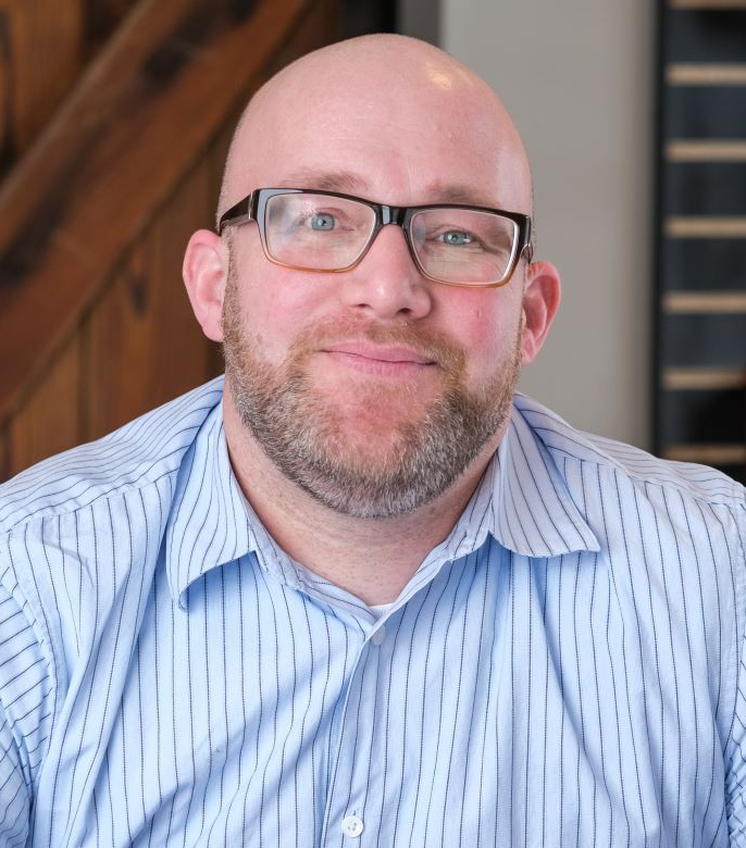 Blonde woman in blue striped shirt and glasses smiles with arms crossed.