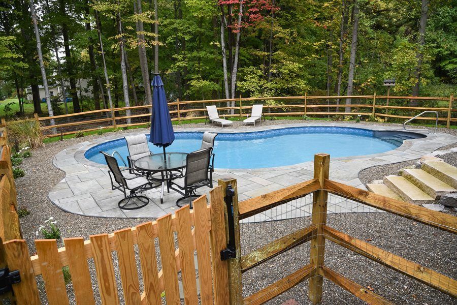 A wooden fence surrounds a large swimming pool with a table and chairs.