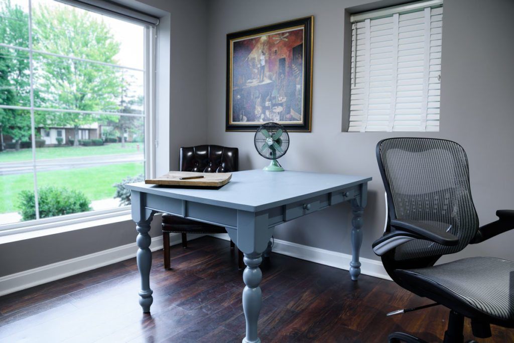 A home office with a desk , chair , fan and a large window.