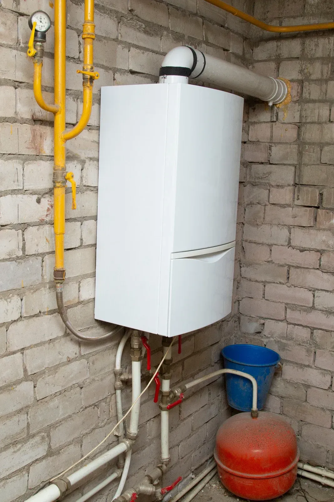 A white boiler is sitting in a brick room next to a blue bucket.