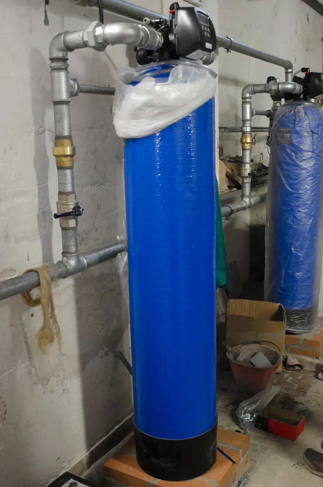 A blue water filter is sitting on top of a brick floor next to pipes.
