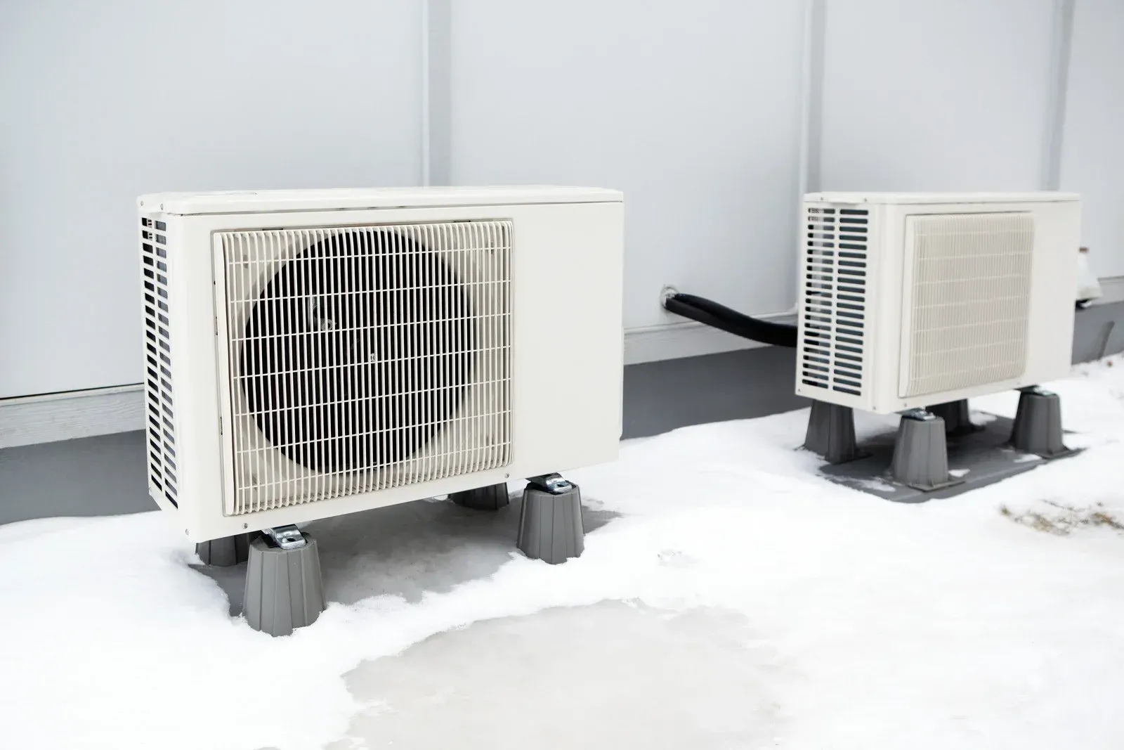 Two air conditioners are sitting in the snow on the side of a building.