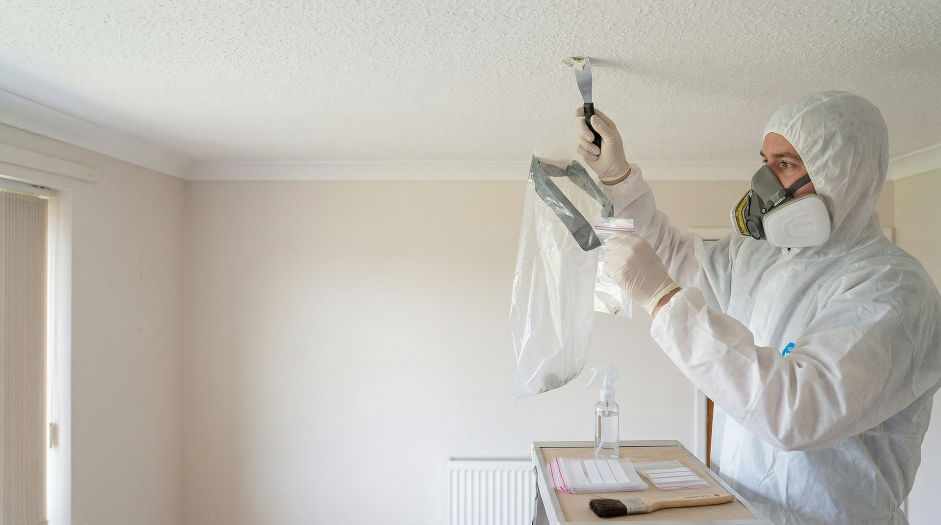 Surveyor in white protective coveralls and respirator taking a controlled asbestos sample from a ceiling in a Dundee home