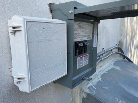 Exterior electrical panel with open cover, mounted on a white wall. Gray box with breakers, conduit. White painted enclosure on the left.