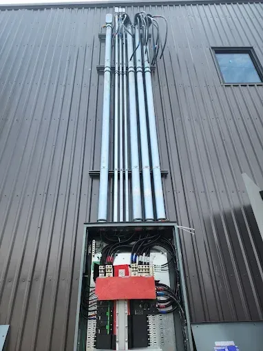 Open electrical panel with bundled conduits running up a corrugated metal wall exterior